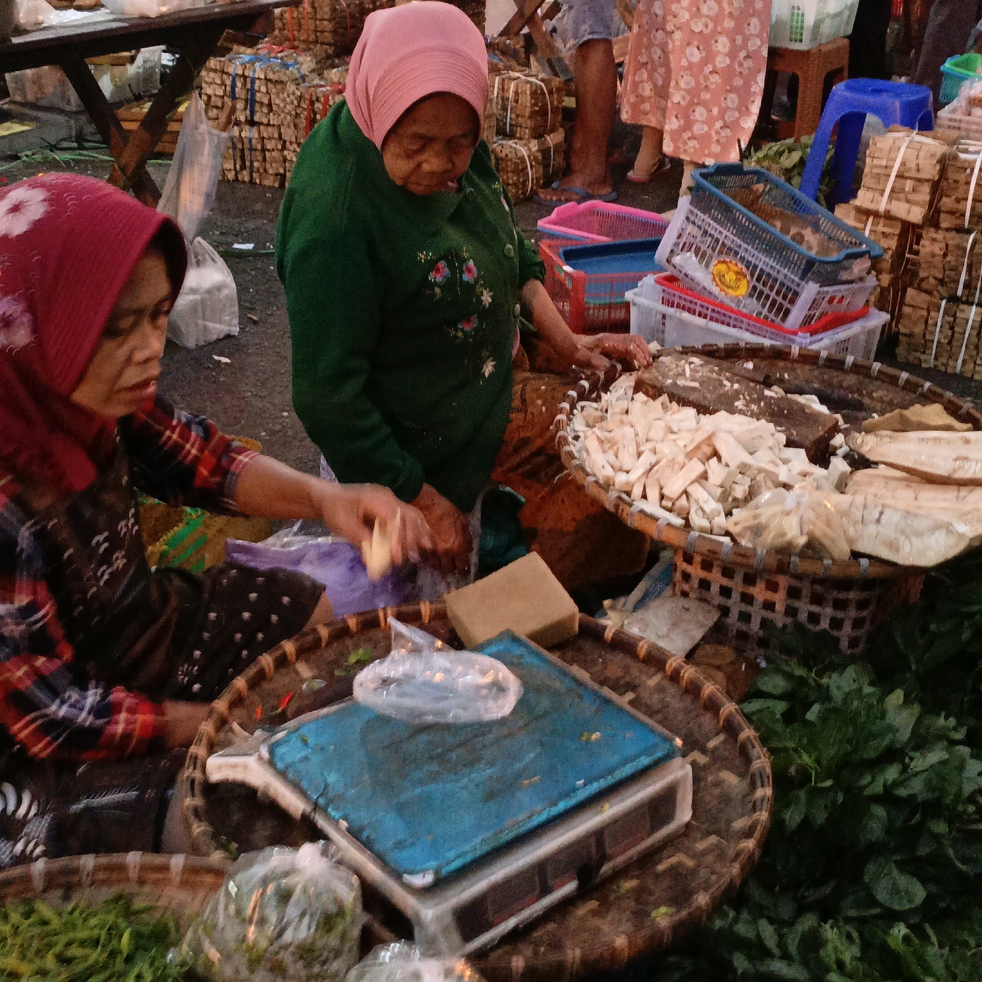 The image depicts a street market scene in Salatiga, Indonesia, likely during a market day. In the foreground, two women are engaged in what appears to be the sale of produce. The woman on the left, wearing a red headscarf and a red and black plaid shirt, is handling some items with her right hand. To her right, an older woman in a green long-sleeved top and a pink headscarf is also interacting with goods, her hands close to a basket filled with chopped food items. A mechanical scale with a blue platform is positioned between them. In the background, stacks of bundled items, possibly dried goods or firewood, are visible, along with plastic crates and baskets. The ground is wet and appears to be paved, typical of an outdoor market. The overall atmosphere suggests a bustling commercial environment.