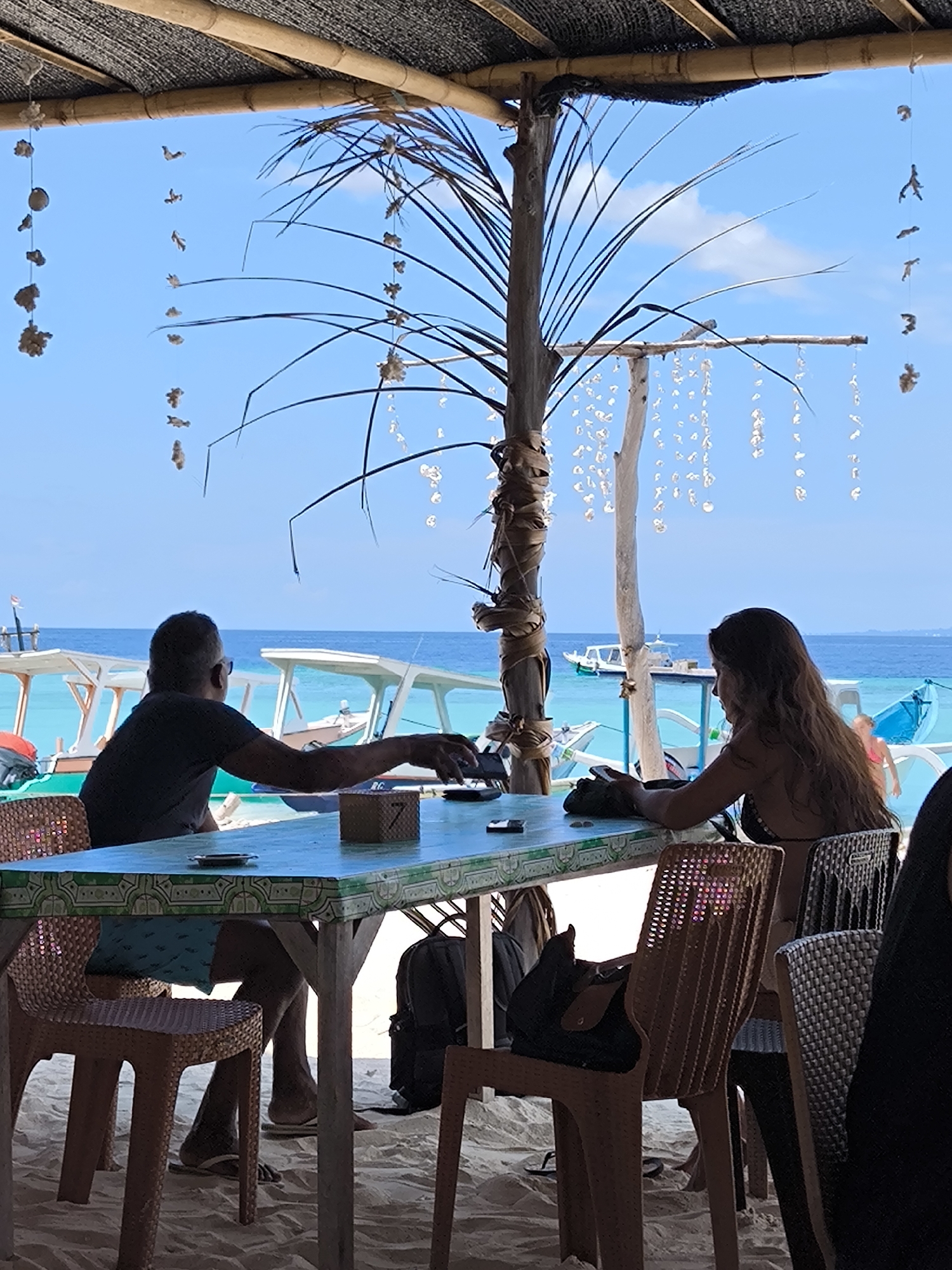 A couple is seated at a blue table, enjoying a view of the ocean. The location is in Unknown, Indonesia. The table is adorned with a number "7" and small items like phones and an ashtray. A tree with hanging shells serves as a decorative element. Boats are visible on the water, suggesting a coastal setting. The environment is sandy, indicating a beach or beachfront establishment.