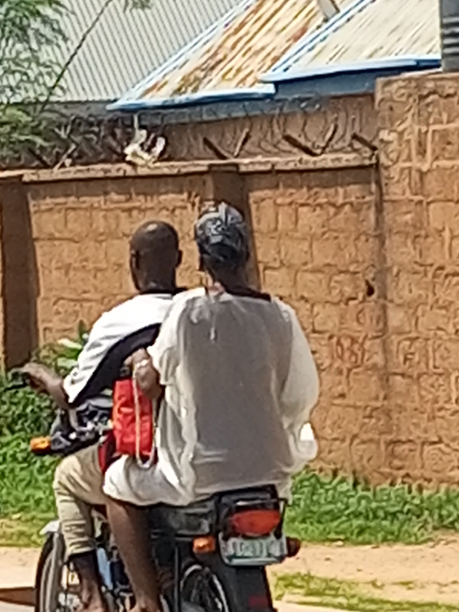 In Tempure, Nigeria, two individuals are observed riding a motorcycle. The person in the rear, wearing a gray t-shirt, is positioned behind the driver, who is dressed in a white t-shirt with black accents. A red bag is slung over the driver's shoulder. The motorcycle has a visible license plate. The background features a textured, light brown brick wall and a sloped corrugated metal roof of a building. Green vegetation is present along the lower left and right sides of the image, with a dirt path in the foreground.