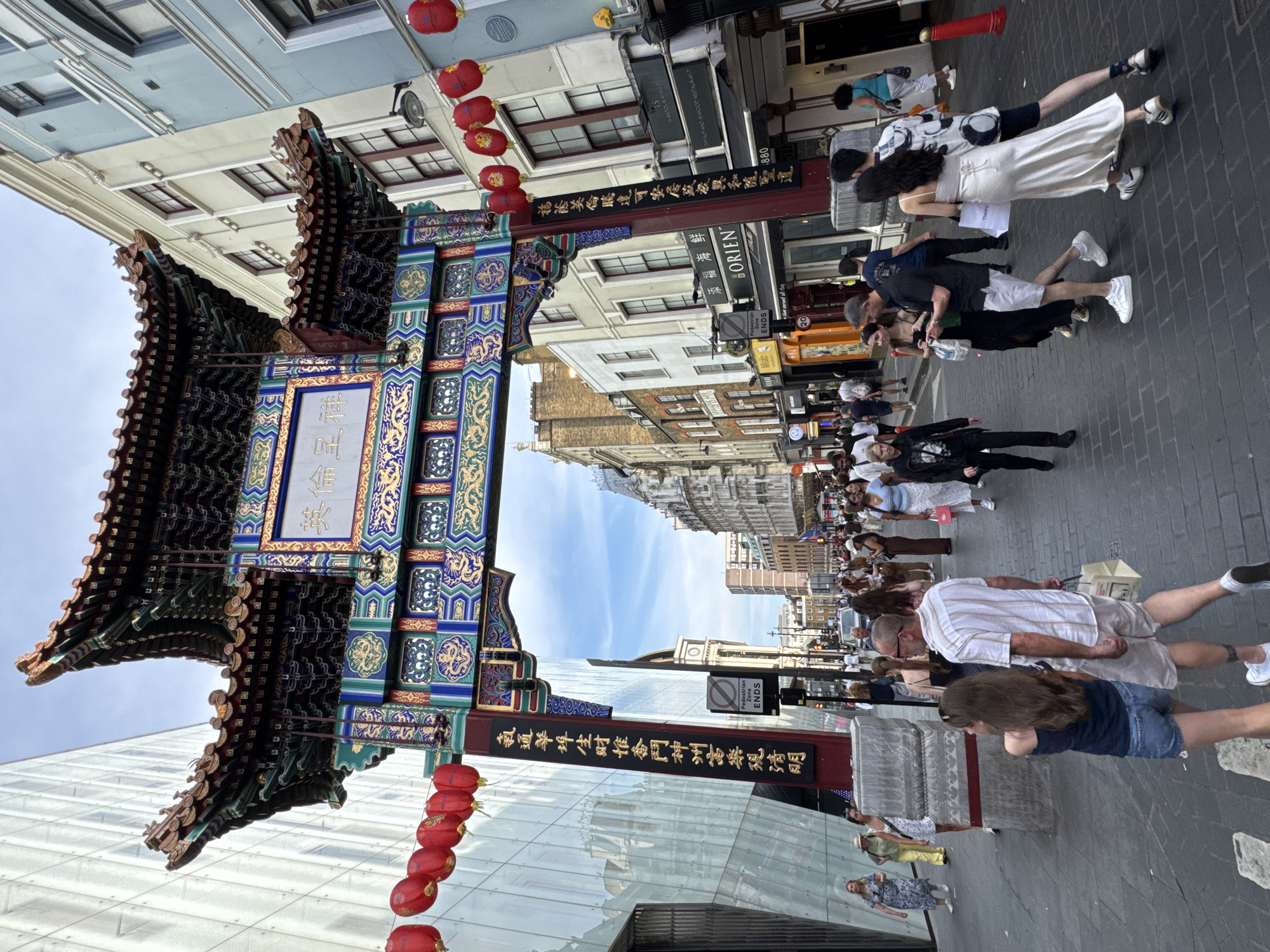 The image depicts a view of Chinatown Gateway in London, United Kingdom. The gateway, a traditional Chinese arch, is elaborately decorated with red, green, and gold ornamentation and features Chinese characters. Red lanterns are strung across the street, enhancing the festive atmosphere. The street is paved with cobblestones and lined with buildings of varying architectural styles, some modern with glass facades and others traditional. A diverse crowd of people are walking along the street, some individually, and some in groups. They are dressed in casual summer attire. The scene captures a vibrant urban environment with a blend of cultural influences.