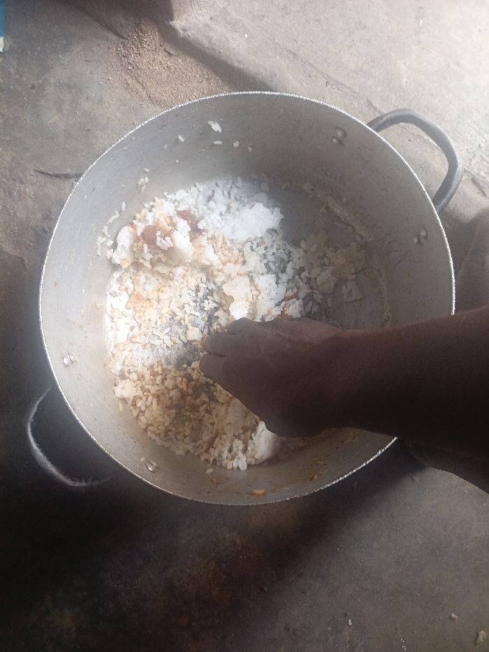 A dark-skinned human hand and forearm are captured in a top-down view, reaching into a large, circular, metallic cooking pot. The hand is open, with fingers splayed, making contact with the pot's contents. The pot holds a mixture of white rice, substantial dark-brown to black burnt rice fragments concentrated at the bottom, and reddish-brown residue, suggesting a recently prepared and partially consumed or mixed meal. The pot features two black handles, one partially visible on the upper right, and rests directly on a rough, textured, grey-brown ground surface, consistent with concrete or packed earth. The scene is located in Unknown, Unknown.