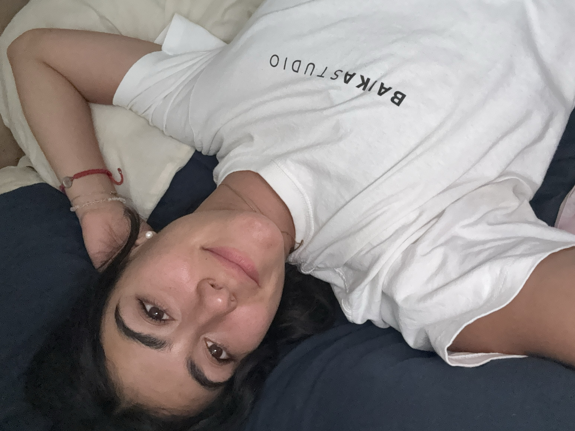 A woman is lying on a bed with navy blue sheets. She is wearing a white t-shirt with "BAIKA STUDIO" printed on it. Her head rests on a navy blue pillow. She is wearing a pearl earring on her left ear and has bracelets on her left wrist. The location of the photo is unknown.