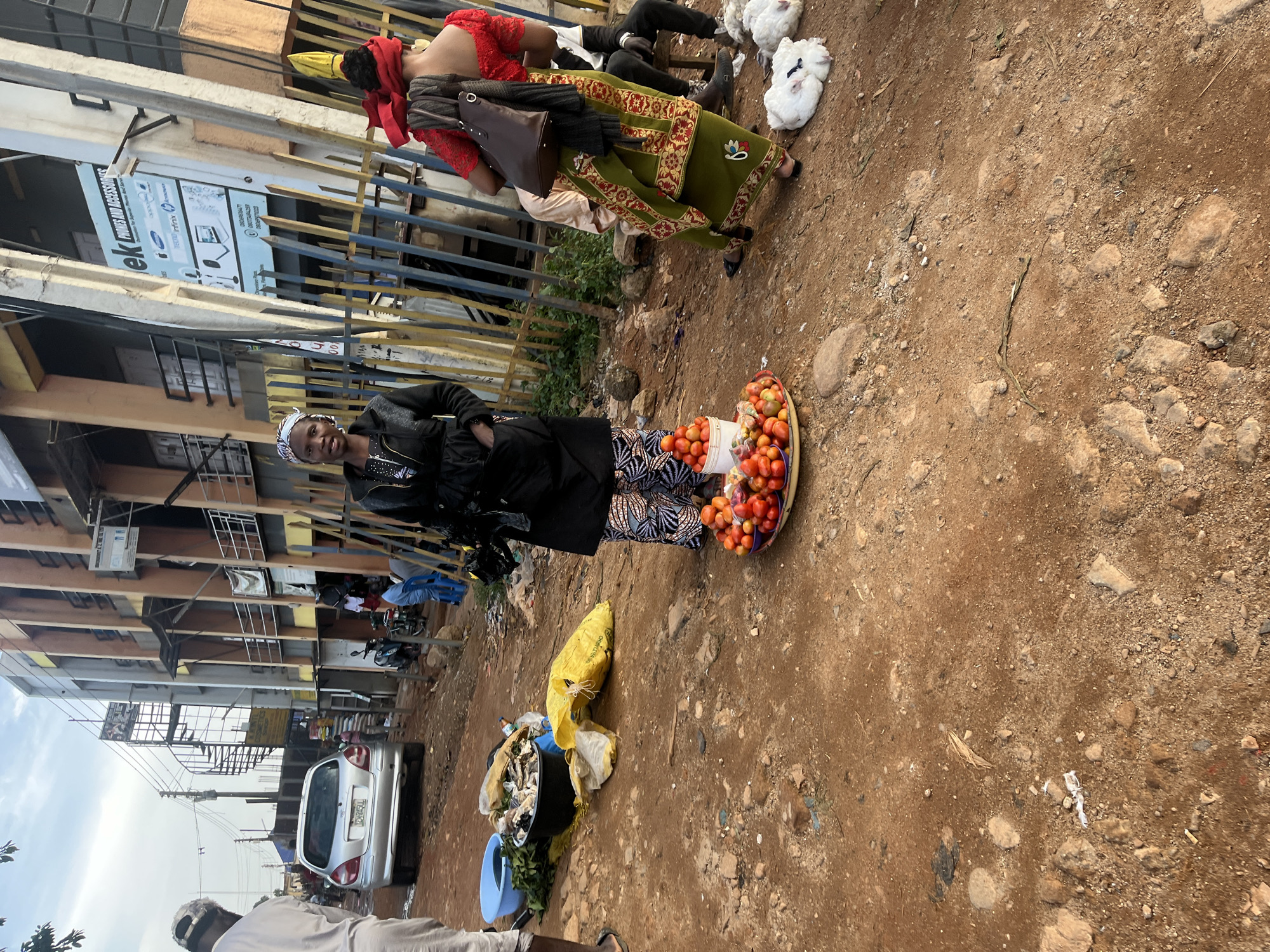 A street scene in Jos, Nigeria, is depicted, featuring a woman standing in the foreground beside a tray of tomatoes. She is wearing a black jacket, patterned pants, and a headscarf. Behind her, a silver car is parked on the dirt road, and buildings with multiple stories line the street. Several other individuals are visible in the background, some walking and others sitting. Two white chickens lie on the ground near the right edge of the frame. The environment appears to be an outdoor market or commercial area with a mix of commercial establishments and informal vendors.