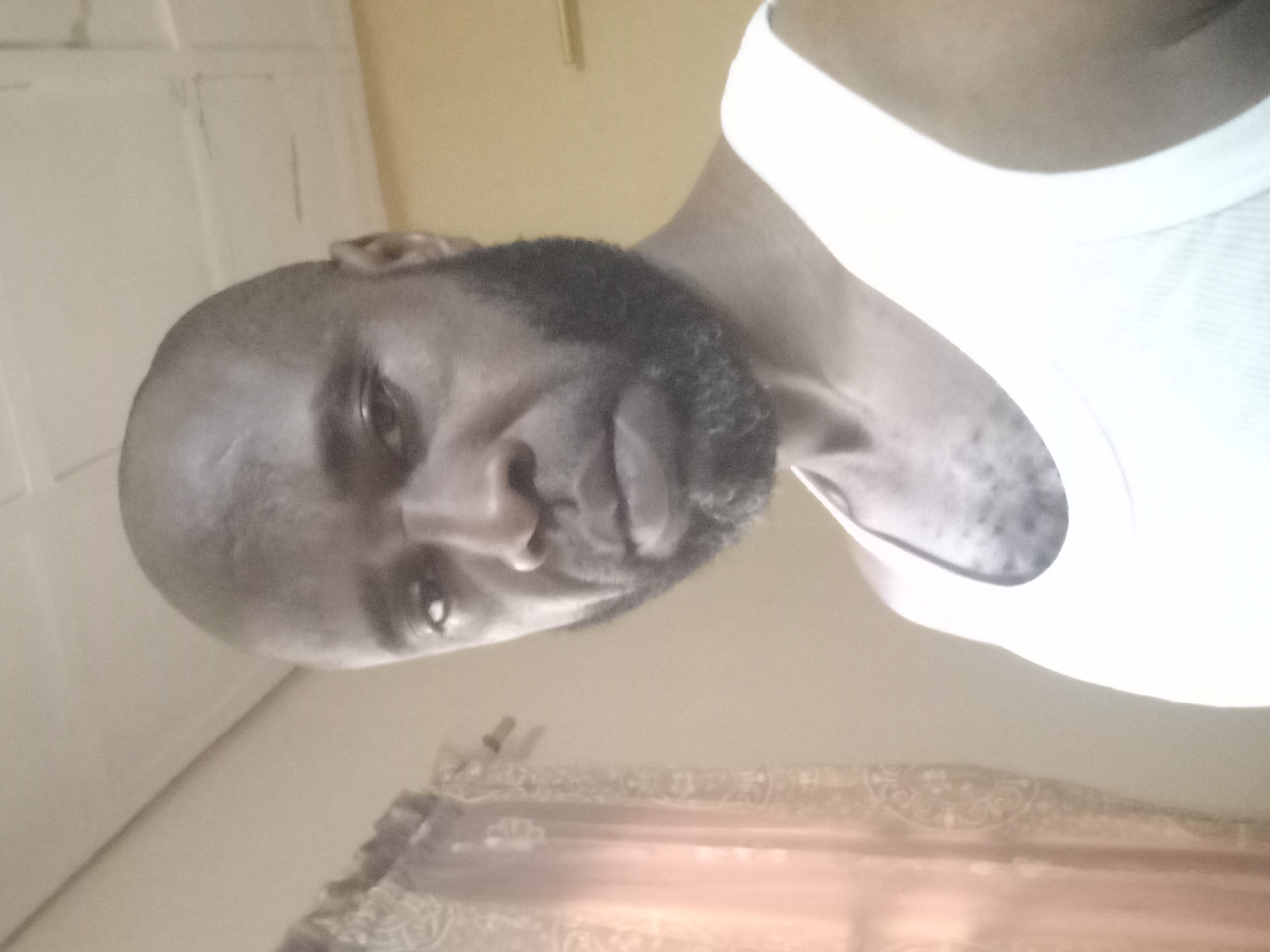 A dark-skinned man with a shaved head and full beard is wearing a white tank top and looking directly at the camera. He is positioned in an indoor setting with a white door visible in the background on the left. A patterned curtain or fabric is visible along the bottom edge of the frame. The location is presumed to be Barden Yana, Nigeria.