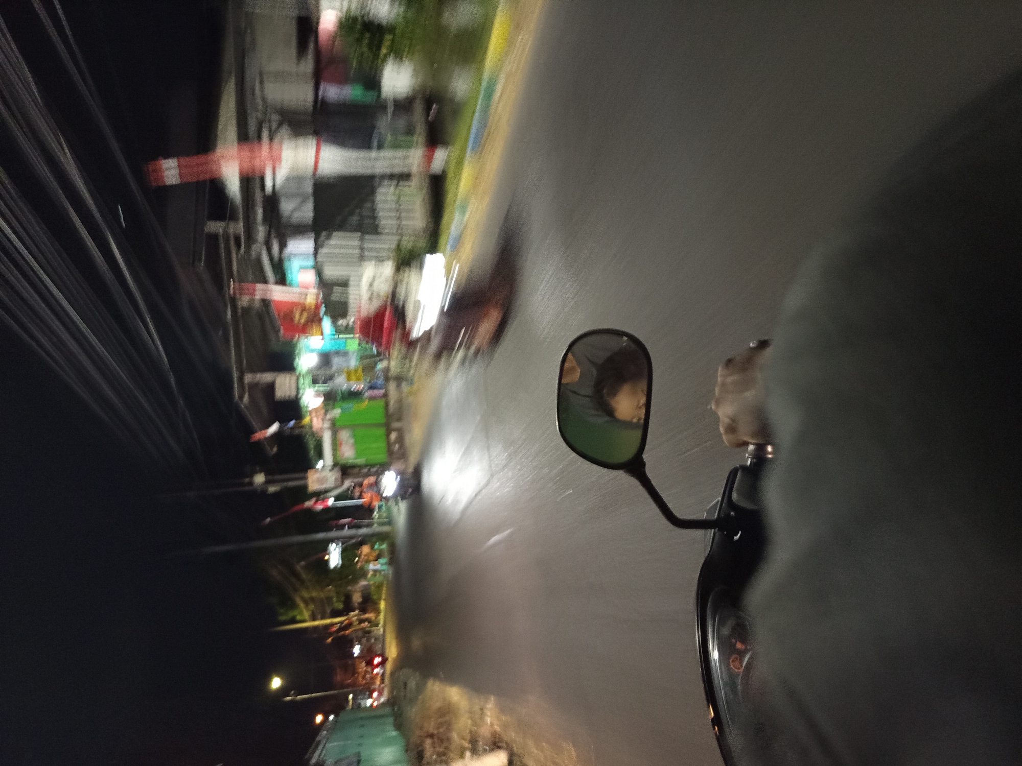 The image captures a nighttime scene on a street in Punggul, Indonesia, from the perspective of someone riding a motorcycle. The road is paved and wet, reflecting the streetlights and the headlights of other vehicles. Along the sides of the road are various buildings, some with illuminated signs, and vegetation. Several motorcycles are visible, some moving and some stationary. The rider's hand is visible on the handlebar, and a side mirror reflects a blurry image of a person's face. The sky is dark with visible power lines overhead. The overall atmosphere suggests a typical urban environment at night, with signs of commercial activity and traffic.