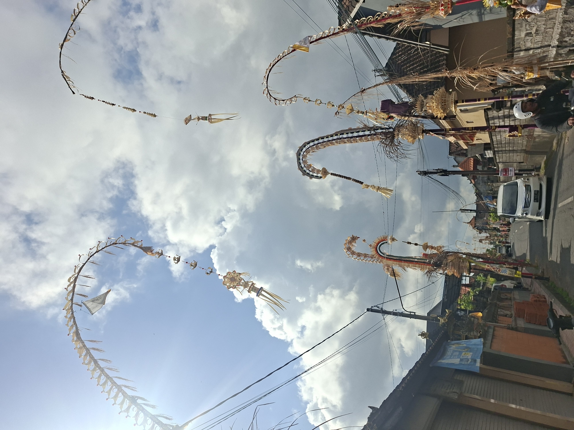 In Blahbatuh, Indonesia, a street is lined with traditional Balinese decorations known as Penjor, which are tall, curved bamboo poles adorned with woven palm leaves, flowers, and offerings. These Penjor are erected for religious ceremonies, particularly during the Galungan and Kuningan festivals. The image captures a bright, sunny day with some clouds, casting shadows on the street and buildings. Several Penjor are visible along the road, arching gracefully over the street. On the right side of the street, a man wearing a white helmet rides a motorcycle. A white van is seen driving down the street, and other vehicles are parked or moving in the background. The buildings along the street appear to be typical Balinese houses and shops, with tiled roofs and intricate architectural details. The overall scene depicts a festive atmosphere, indicative of a cultural celebration in Bali.
