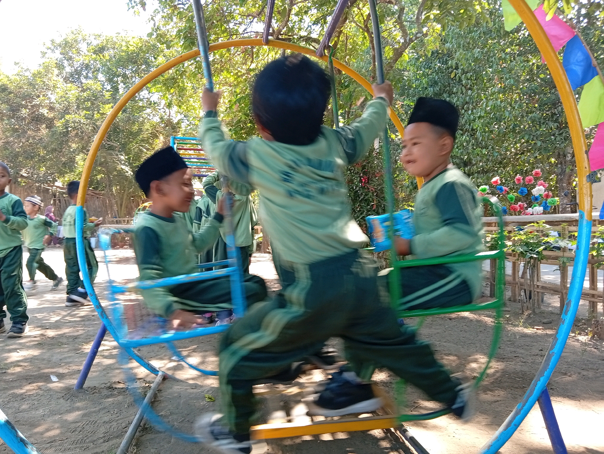 This image captures a lively scene at a playground in Miritpetikusan, Indonesia. A group of young children, dressed in matching green uniforms and black caps, are actively playing on a circular swing set. The swing set features blue, yellow, and green accents, with colorful flags adorning the top. Several children are in motion, with their figures blurred due to the movement. One child in the foreground, seen from behind, is on the swing, their arms extended. Another child to the right, facing the viewer, is smiling and also on a swing. Other children can be seen in the background, also wearing similar uniforms. The ground beneath the playground is sandy and dirt-covered, with sparse vegetation and trees providing a natural backdrop. The sunlight suggests it is daytime.