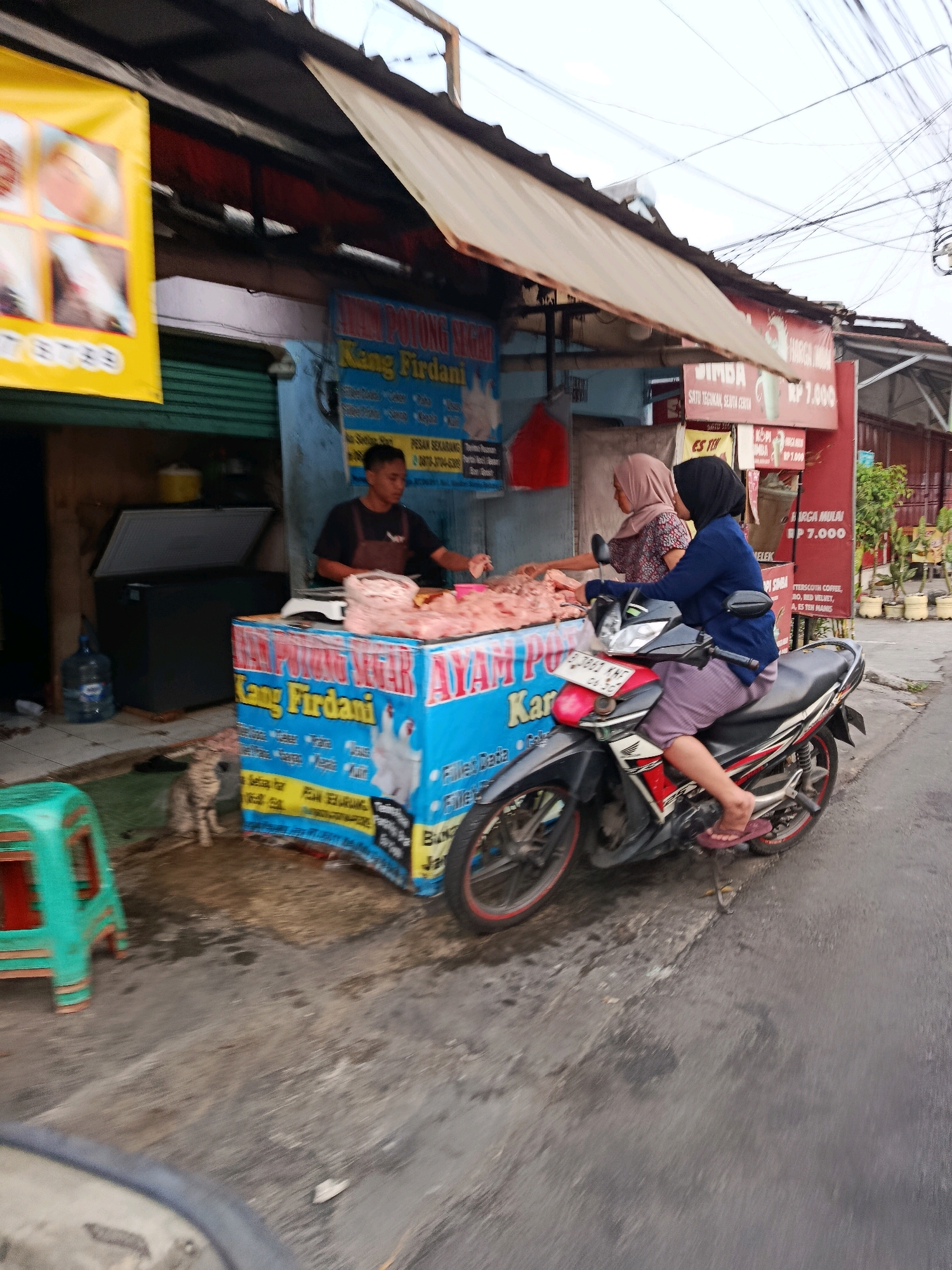 The image depicts a street scene in Bekasi, Indonesia, focusing on a meat stall. A vendor, a young man wearing a dark apron over a black shirt, stands behind a blue and yellow tarp-covered stall displaying cuts of meat. Two women are present. One, wearing a light brown hijab and a patterned shirt, appears to be a customer, interacting with the vendor. The second woman, wearing a black hijab and a dark blue long-sleeved shirt, is seated on a motorcycle parked on the street, facing away from the stall, and appears to be waiting. The motorcycle is a red, white, and black Honda. A grey cat is visible on the ground near the stall. Behind the stall, a large, dark freezer with its lid open is present, along with a blue water jug. A green plastic stool is positioned to the left of the frame. Various signs are visible, including a yellow banner with images on the left and red and blue signs behind the stall with Indonesian text, one of which mentions "KANG FIRDANI" and prices. The environment appears to be an outdoor market or roadside area with asphalt paving and overhead electrical wires.