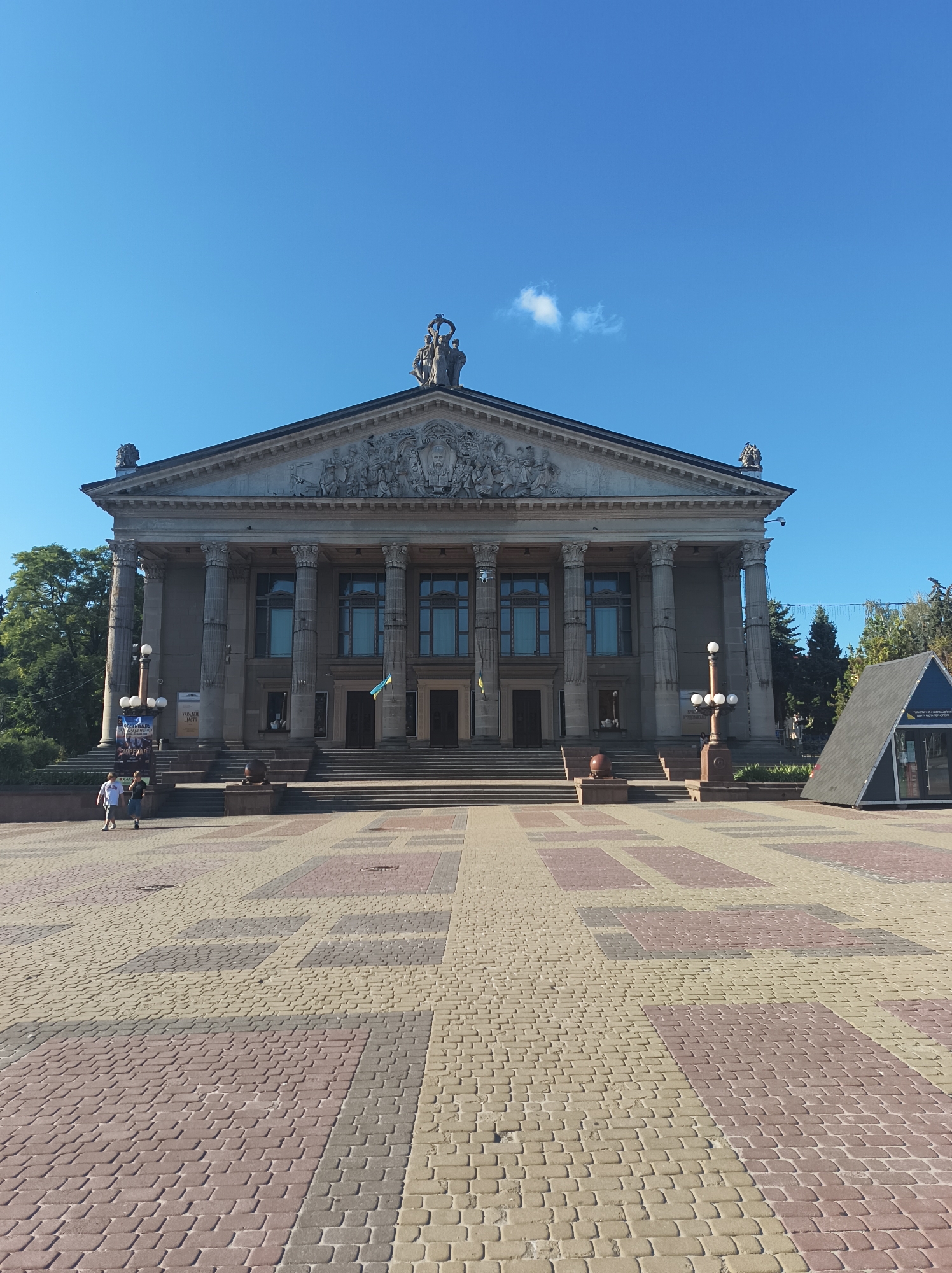 The image shows a wide, paved plaza in front of a large, classical-style building in Ternopil, Ukraine. The building features a grand facade with numerous columns, a pediment adorned with sculptures, and tall windows. Two Ukrainian flags are visible hanging from the building. In the foreground, the plaza is paved with interlocking bricks in a pattern of light beige and reddish-brown. Two individuals, one in a white t-shirt and the other in a dark t-shirt and jeans, are walking away from the camera towards the building. To the right of the building, a modern, dark-colored triangular structure with glass panels is situated on the plaza. The sky is a clear, bright blue with a few wispy clouds. Trees are visible behind the building on the left and on the right. The overall scene depicts a public space outside a significant architectural landmark.