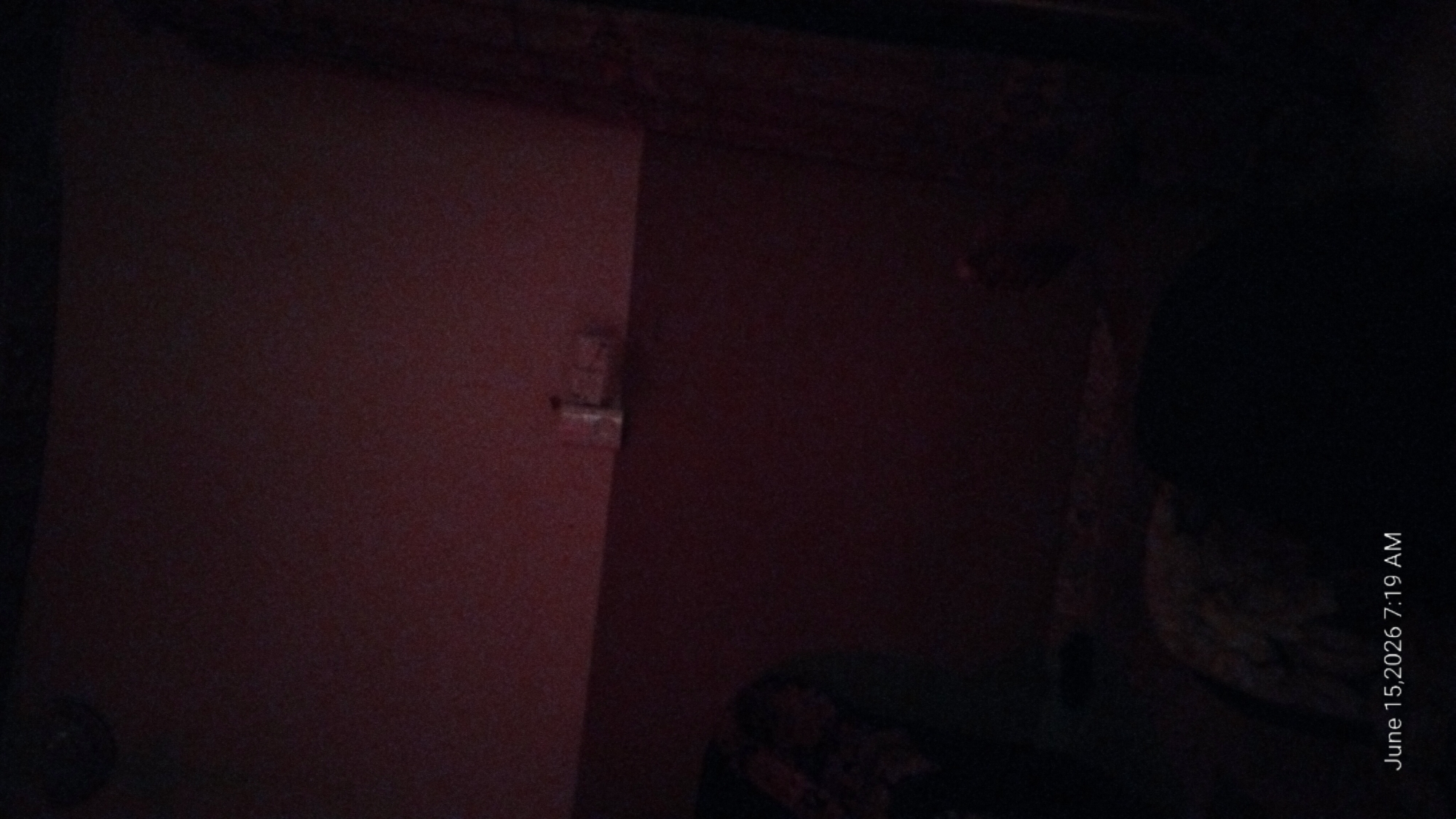The image depicts a dimly lit room. In the upper right quadrant, a white object, possibly a light switch or an outlet, is visible on the wall. The lower portion of the image is almost entirely black, suggesting very low light conditions. The timestamp "June 15, 2026 7:19 AM" is overlaid in the bottom left corner. The location in Howayi, Nigeria, is not ascertainable from the visual information presented in the image. There are no discernible people, specific objects, or actions that can be identified with certainty.
