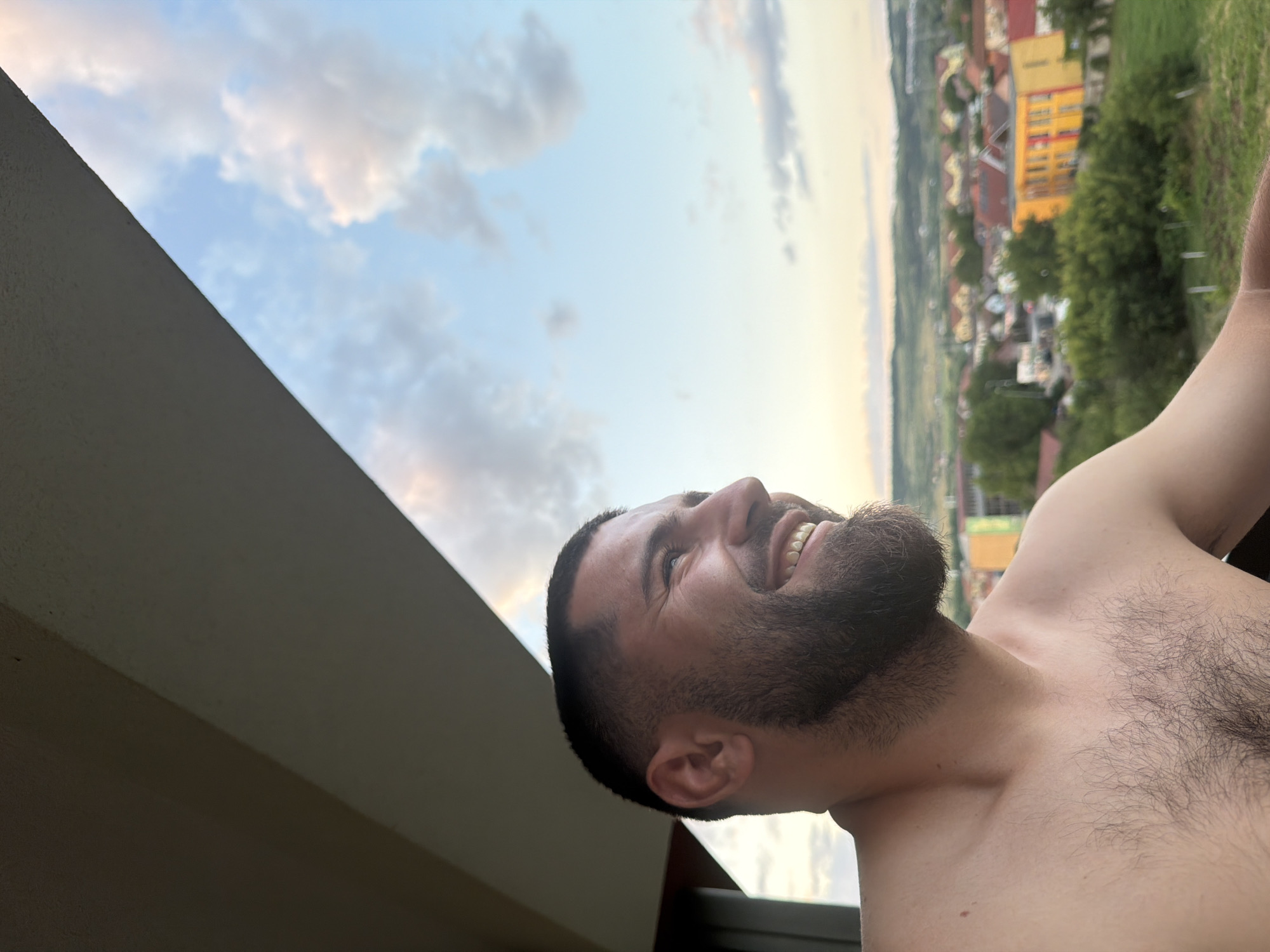 The photograph captures a close-up, chest-up view of a shirtless man with a beard, looking to his right and smiling. He is positioned on a balcony, as indicated by the overhead structure and railing visible in the foreground. The background reveals a sprawling town or village under a partly cloudy sky, suggesting an outdoor setting. The architecture of the buildings appears to be residential, with a mix of red-tiled roofs and brightly colored facades, one of which is yellow and red. Lush green trees and vegetation dot the landscape, contributing to a serene, possibly rural or suburban, atmosphere. The lighting suggests either dawn or dusk, with soft, warm tones bathing the scene. This scene is likely located in Florești, Romania, a commune known for its residential development and proximity to natural landscapes.