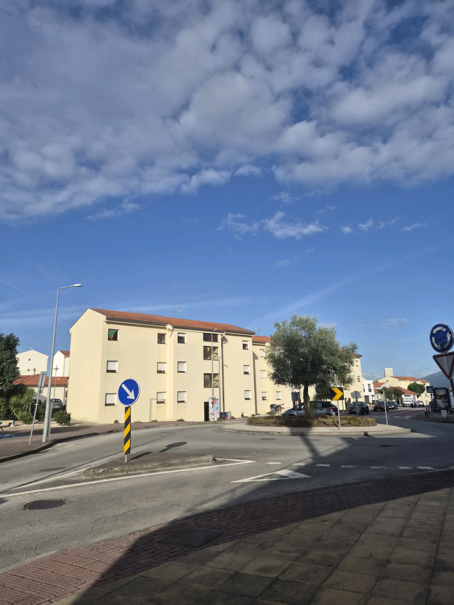 The image captures a street scene in Barcelos, Portugal, with a residential building dominating the background. The building is painted a light yellow with a red-tiled roof and features multiple windows with white shutters. A Portuguese flag is visible in one of the windows. In the foreground, a roundabout is depicted with traffic signs, including a circular sign with a blue background and a white arrow indicating the direction of travel, and a yellow directional arrow sign. A tree with a rounded canopy stands in the center of the roundabout. Several cars are present on the road and around the roundabout. The sky is blue with scattered white clouds. The sidewalk in the foreground is paved with reddish-brown brick and gray concrete tiles, with a shadow cast across it. A street lamp is also visible on the left side of the image.