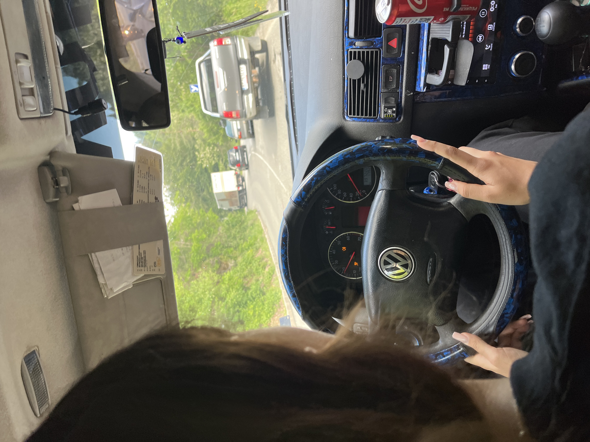 The image shows the interior of a Volkswagen car, viewed from the passenger's perspective. A person, only partially visible, is driving. The dashboard features a steering wheel with a Volkswagen logo, a speedometer, and various controls. Decorative blue trim is visible on the steering wheel and dashboard. Outside the car, a road is visible with other vehicles, including a truck and a pickup truck, and trees lining the roadside, suggesting a rural or suburban setting. A can of Coca-Cola is placed in a cup holder. The location is identified as Vlahovo, Bulgaria.