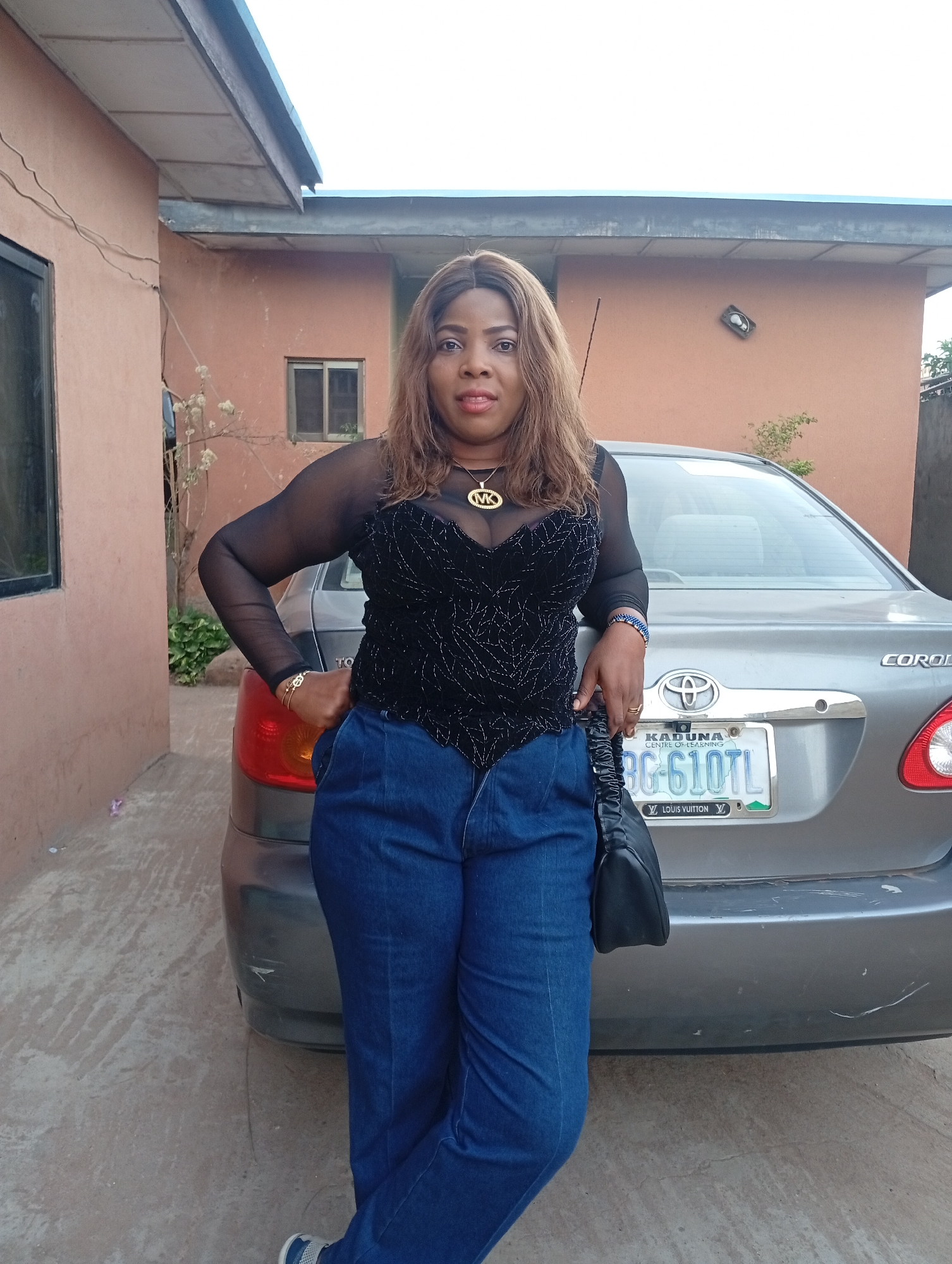 A middle-aged woman stands next to a silver Toyota Corolla sedan. The woman has brown hair and wears a black sheer-sleeved top, dark blue jeans, and sneakers. She has jewelry on her neck, wrist, and fingers, and a black purse hangs from her shoulder. The car has a license plate that reads "KADUNA CENTRE OF LEARNING BG-610TL LOUIS VUITTON." The backdrop includes a peach-colored building with a small window, a larger window, and green vegetation. The location is in Sabon Tasha, Nigeria.