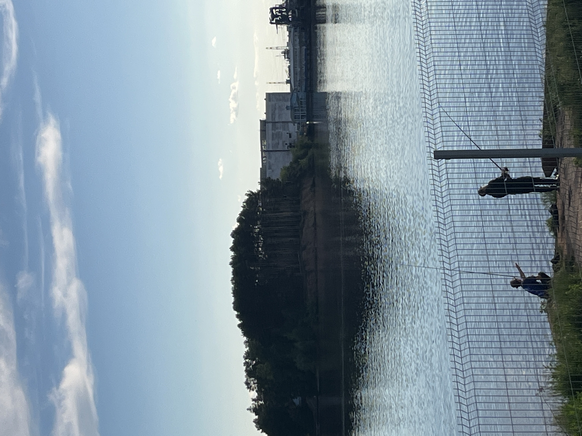 The image depicts a serene daytime scene, likely along a wide body of water in Chaikovsky, Russia. Two individuals are engaged in fishing from a paved bank or pier, separated from the water by a prominent wire mesh fence.
In the foreground, on the right side of the image, two figures are visible. One, wearing dark clothing, stands closer to the viewer, actively holding a fishing rod that extends into the calm water. Further along the bank, a second person, dressed in a blue top, is also fishing with a rod cast out. The wire mesh fence runs horizontally across the lower and middle right of the image, featuring a distinct grid pattern.
The majority of the image is dominated by the expansive surface of a river or lake, showing subtle ripples and reflections of the sky and distant land.
On the far bank, a dense line of dark green trees creates a natural silhouette against the lighter sky, suggesting a wooded shoreline or elevated land. Beyond the trees, in the distant background, an industrial landscape comes into view, characterized by several large, light-colored buildings and what appear to be cranes or heavy machinery, indicating a port or manufacturing facility.
The sky above is a clear, light blue with scattered, wispy white clouds, suggesting fair weather. The lighting is bright and even, consistent with daytime conditions. There is no visible text in the media file. The overall impression is one of peaceful recreation set against an industrial backdrop.