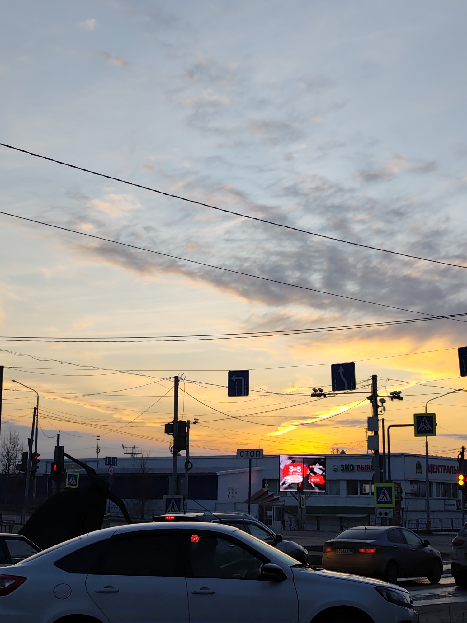 The image shows a street scene in the city of Chaikovsky, Russia at sunset. In the foreground, there are several cars, including a white sedan, stopped at a traffic light. The traffic light is red. Street signs, including pedestrian crossing signs, are visible. In the background, a building with a large digital advertisement screen is visible. The screen displays the words "3=5" in red. The sky is orange and cloudy. Power lines crisscross the sky above the street.