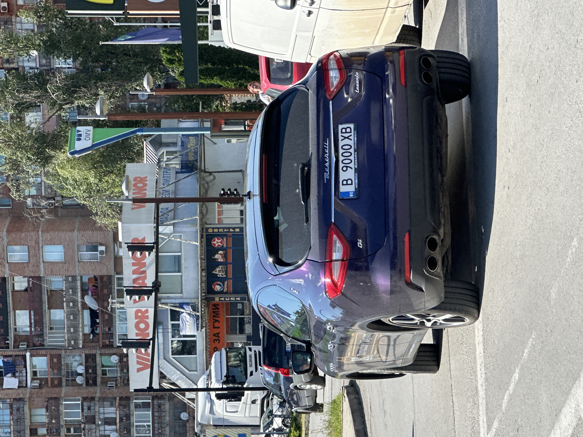 In Veliko Tarnovo, Bulgaria, a dark purple Maserati Levante is parked on a paved road, facing away from the camera. The car's license plate reads "B 9000 XB". Behind the Maserati, a white van and a red car are visible, with a person standing near the red car. Further in the background, a large, multi-story brick building with numerous windows and balconies dominates the scene, along with trees and other commercial structures. Traffic lights and OMV signage are also present. The overall environment suggests an urban setting.