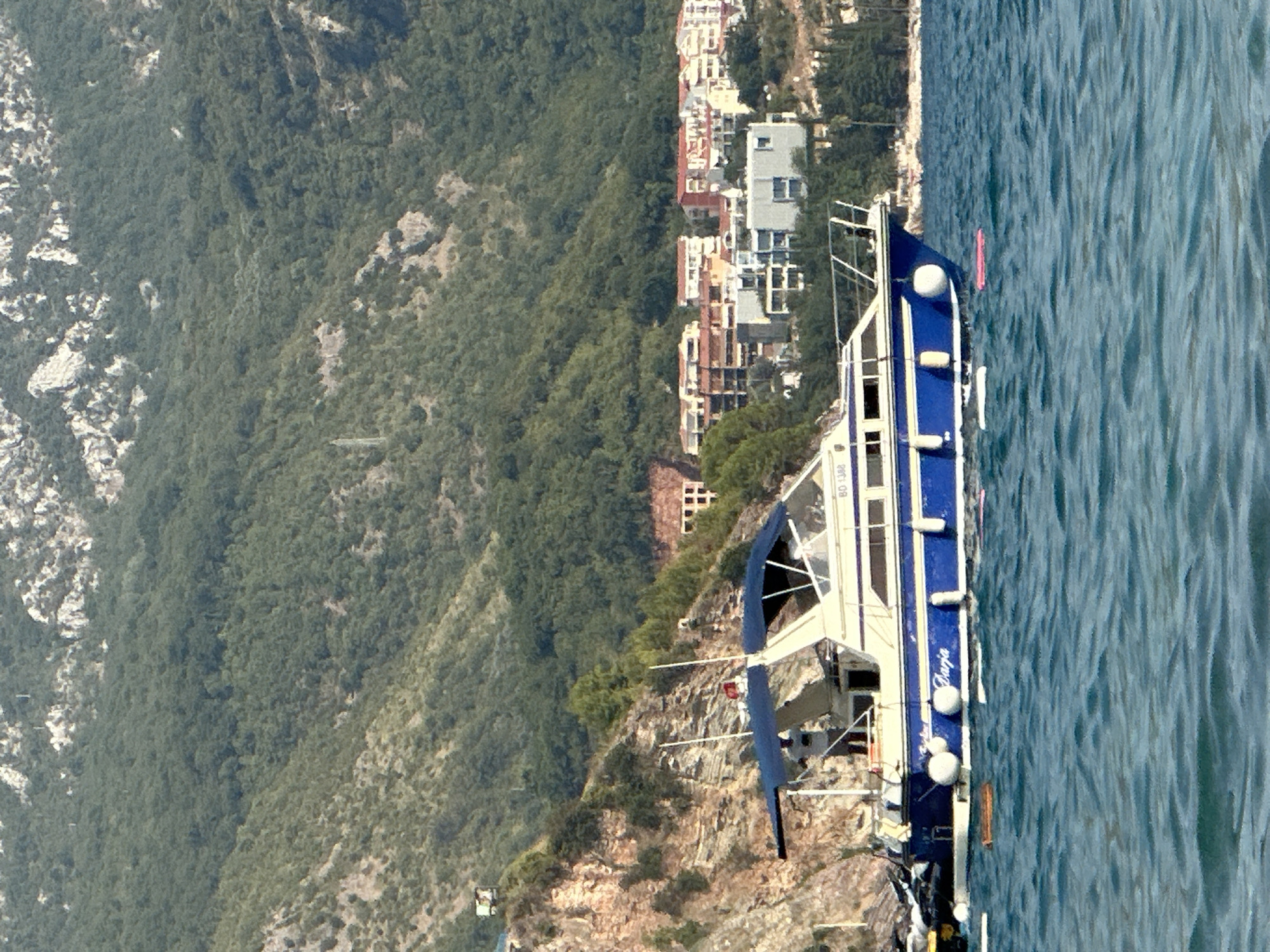 The image shows a blue and white motor yacht anchored in the bay of Boreti, Montenegro. The yacht has a blue canopy over the cockpit and a white cabin with blue trim. The name "Darja" is written in white on the blue hull. Several white buoys are attached to the side of the yacht, and a few inflatable floats are visible in the water around it. In the background, a hillside is covered with lush green trees and rocky outcrops. Several buildings, appearing to be houses or hotels with red-tiled roofs, are nestled into the hillside. The sea is calm with gentle ripples, and the sky is clear and blue. The scene conveys a sense of leisure and tranquility on a sunny day.
