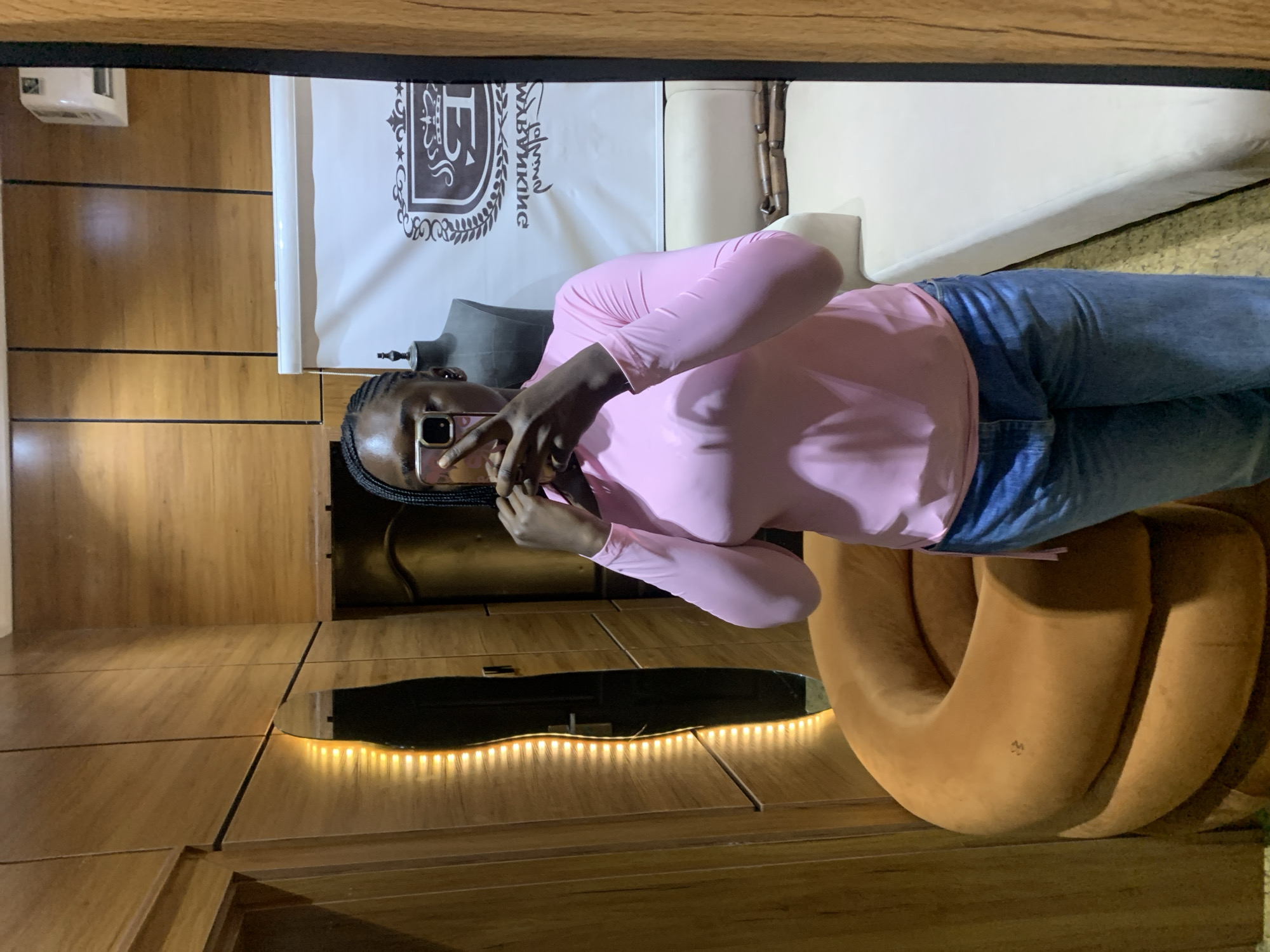 A young Black woman with braided hair is taking a mirror selfie in what appears to be a fitting room or boutique. She is wearing a pink long-sleeved shirt and blue jeans. In the background, a mannequin is visible, along with a couch and a wall decorated with wooden panels and a subtle lighting feature. The overall setting suggests an indoor retail or dressing space.