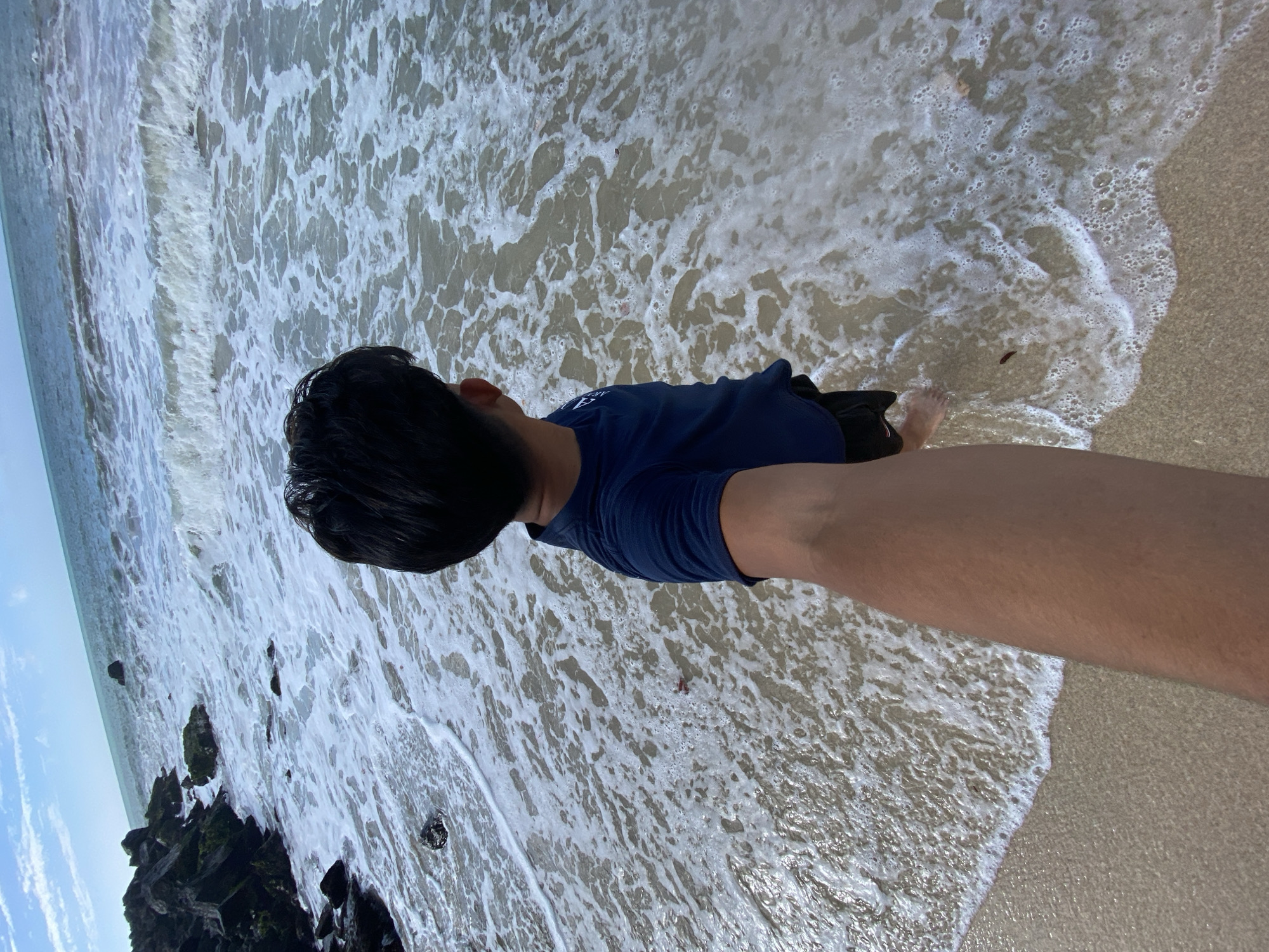 The image shows a person standing at the edge of the ocean on a sandy beach, likely in Pedro González, Venezuela, based on the location request. The person is wearing a dark blue shirt and dark shorts. The waves are washing onto the shore, and some rocks are visible at the edge of the water on the left. The sky is partially visible with scattered clouds. The camera angle appears to be a first-person view, with the person's arm visible in the lower right of the frame.