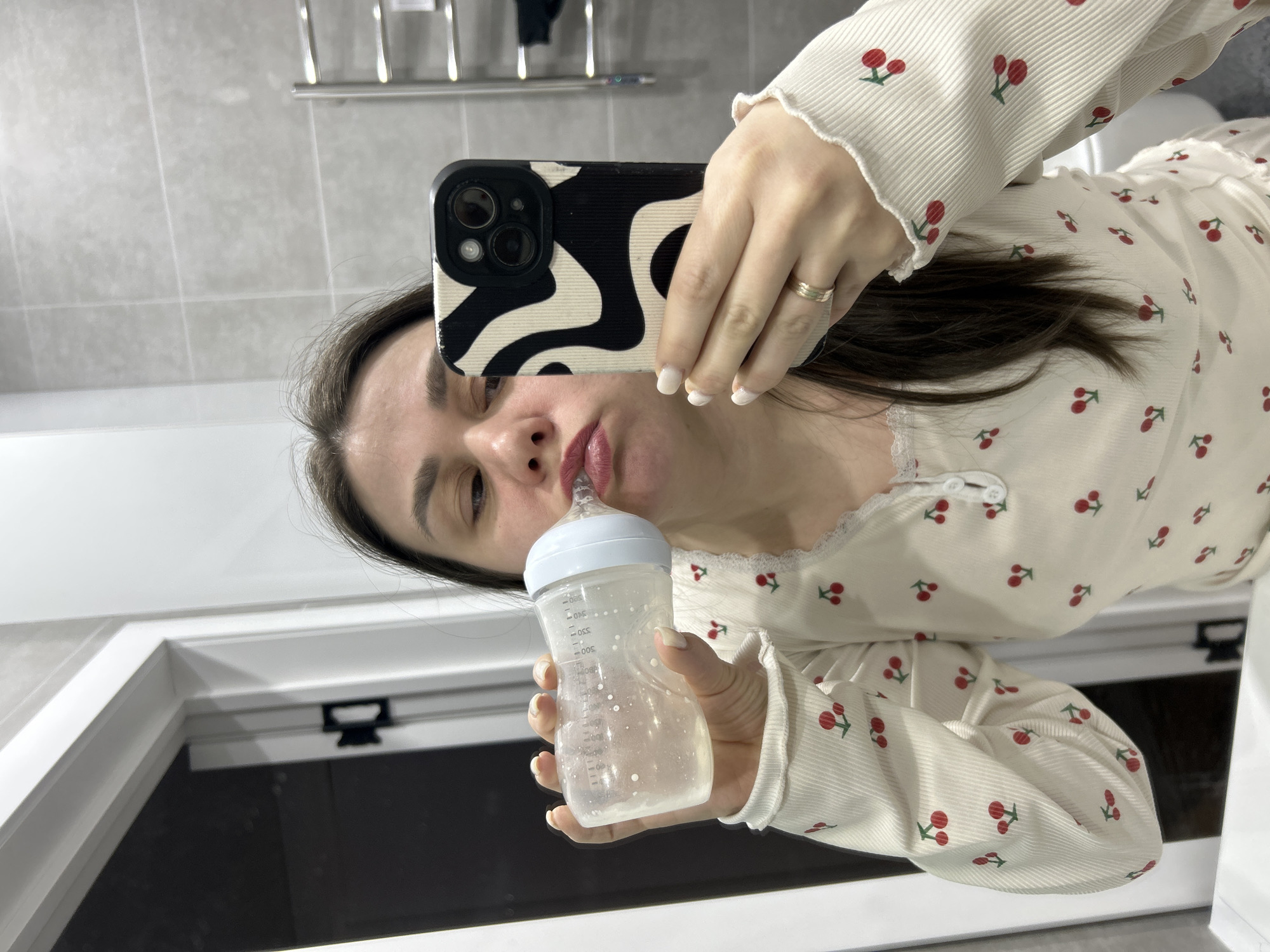 A woman is taking a selfie in a bathroom mirror. She is holding a baby bottle to her mouth with her left hand and a phone with a black and white patterned case in her right hand. She is wearing a light-colored shirt with a cherry print. The bathroom has gray tiled walls and a white countertop with a window or mirror above it. There is a towel rack with a dark-colored towel hanging on it in the background. No definitive landmarks are visible to confirm the location as Chaikovsky, Russia.