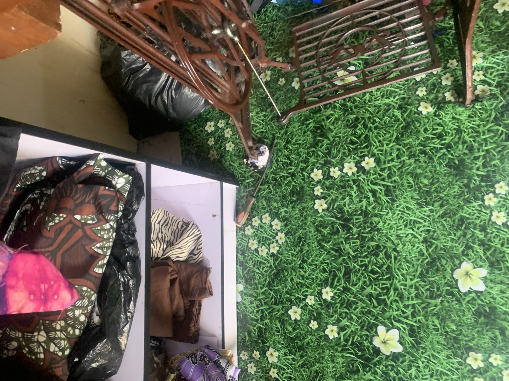 The image depicts an indoor scene, likely a room, with a focus on a sewing machine and various fabrics. A vintage, ornate sewing machine made of dark metal is positioned on the right side of the frame, its base and a portion of the sewing table visible. The floor is covered with a green, artificial grass-like material adorned with scattered white and yellow flowers, creating a whimsical or decorative effect.
To the left of the sewing machine, a white shelf unit is partially visible. On the top shelf, several pieces of fabric are stored, including one with a bold brown and green patterned design and a bright pink fabric piece. Below this, on the lower shelf, more fabrics are arranged, featuring brown and a black and white zebra-striped material. A black plastic bag is also present on the top shelf.
The background behind the shelf unit is a plain, light-colored wall. The overall impression is of a crafting or storage area. The specific location within Jauro Yinu, Nigeria, is not discernible from the visual elements of the image.