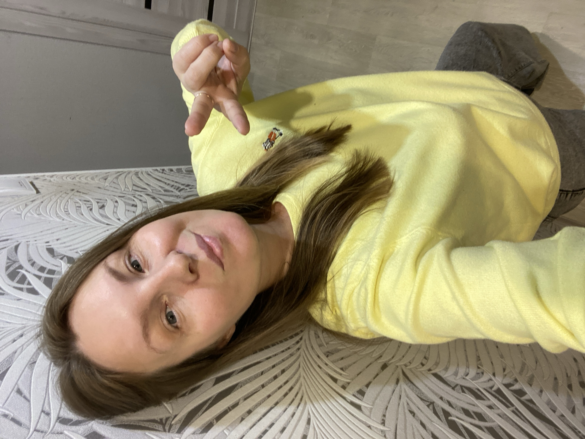 A person with light skin, shoulder-length brown hair, wearing a yellow long-sleeved sweatshirt with a small animal logo on the chest, and gray pants is lying on a surface with a patterned, possibly leafy design. The person is holding up their left hand, which has a ring on the ring finger. The background includes what appears to be a door or wall. There is no evidence of the location being in the city of Noviy, Russia.