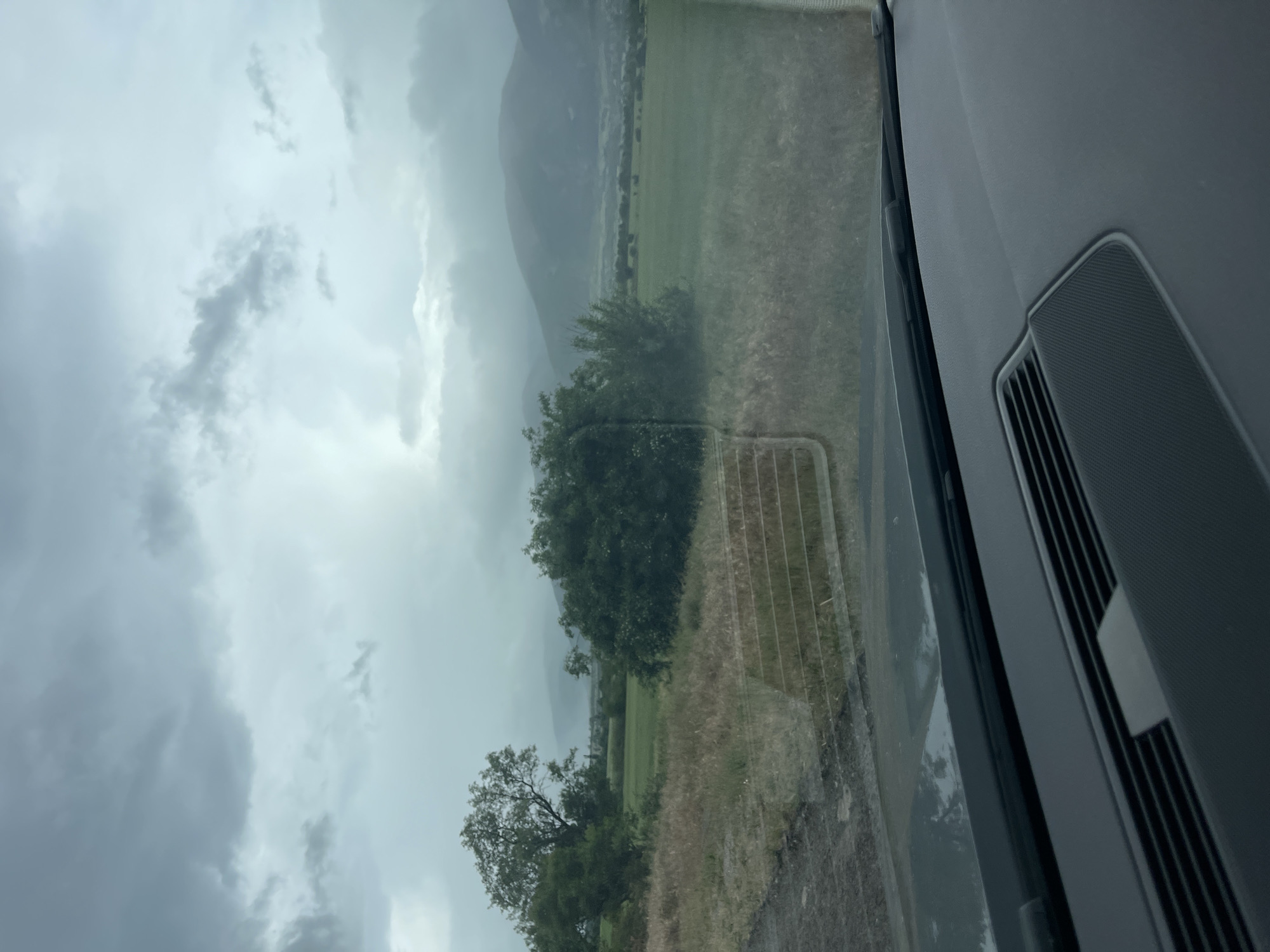 The image, taken from inside a vehicle, shows an overcast scene of a field and mountains near Shivachevo, Bulgaria. The foreground consists of a grassy field with scattered trees, separated by a metal gate or fence. In the distance, there are mountains partially obscured by the cloudy, gray sky. The interior of the vehicle is visible on the right side of the frame, showing a portion of the dashboard.