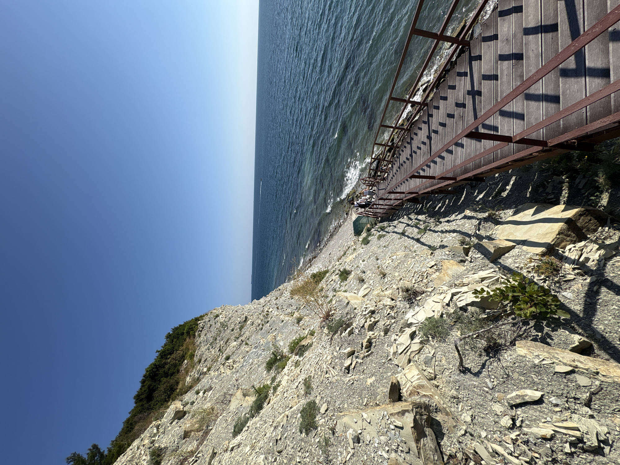 This is a high-angle, wide shot of a wooden staircase descending a rocky cliff face towards the sea, located in Kabardinka, Russia. The sky is a clear, deep blue with no clouds. The rocky cliff to the left is composed of layered gray and beige stone, with sparse vegetation growing from its crevices. The staircase itself is made of weathered wooden planks with dark metal railings on both sides. It is bathed in sunlight, casting distinct striped shadows across the steps. Several people, appearing as small figures from this vantage point, are visible walking down the staircase. The sea is a vibrant blue, with gentle waves lapping at a pebble beach at the base of the cliff. A thin line of white foam marks the edge of the water. In the distance, a small white boat is visible on the horizon.