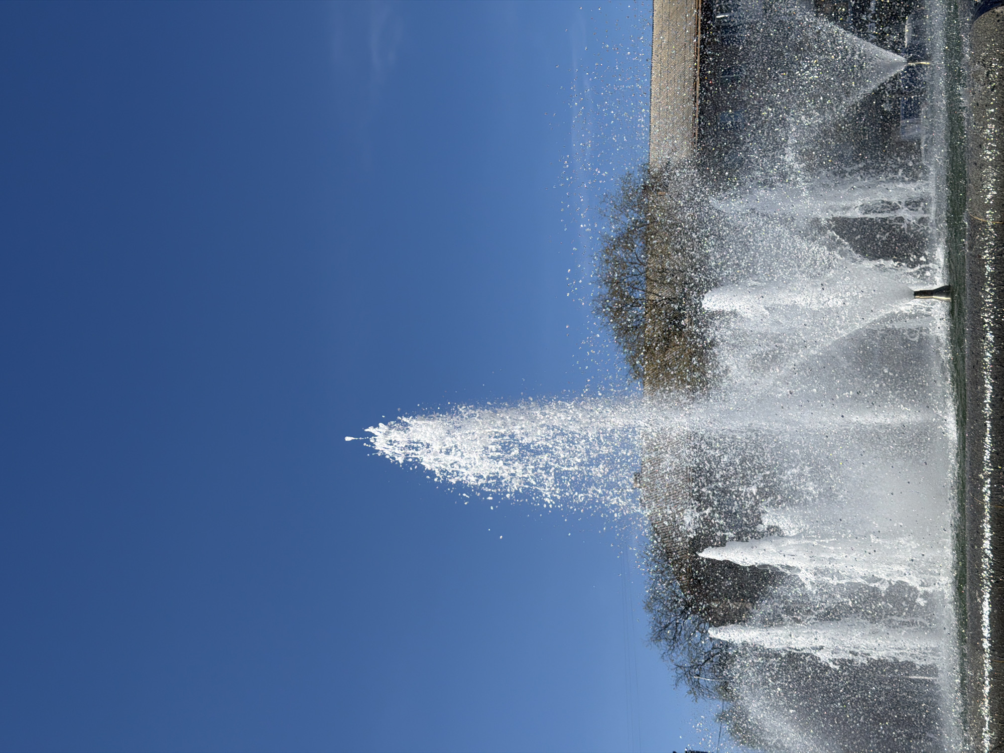 A waterscape in Chaikovsky, Russia is captured on a clear day under a bright blue sky. The image focuses on water jets spraying upwards from a fountain, creating a sparkling, airy effect. Some jets project high into the sky while others spread outwards. The edge of the fountain is made of a dark grey material, showing small reflections. Behind the fountain, a building is partially visible, with bare branches of a tree in the background. The image is composed with the fountain angled, presenting a dynamic view.
