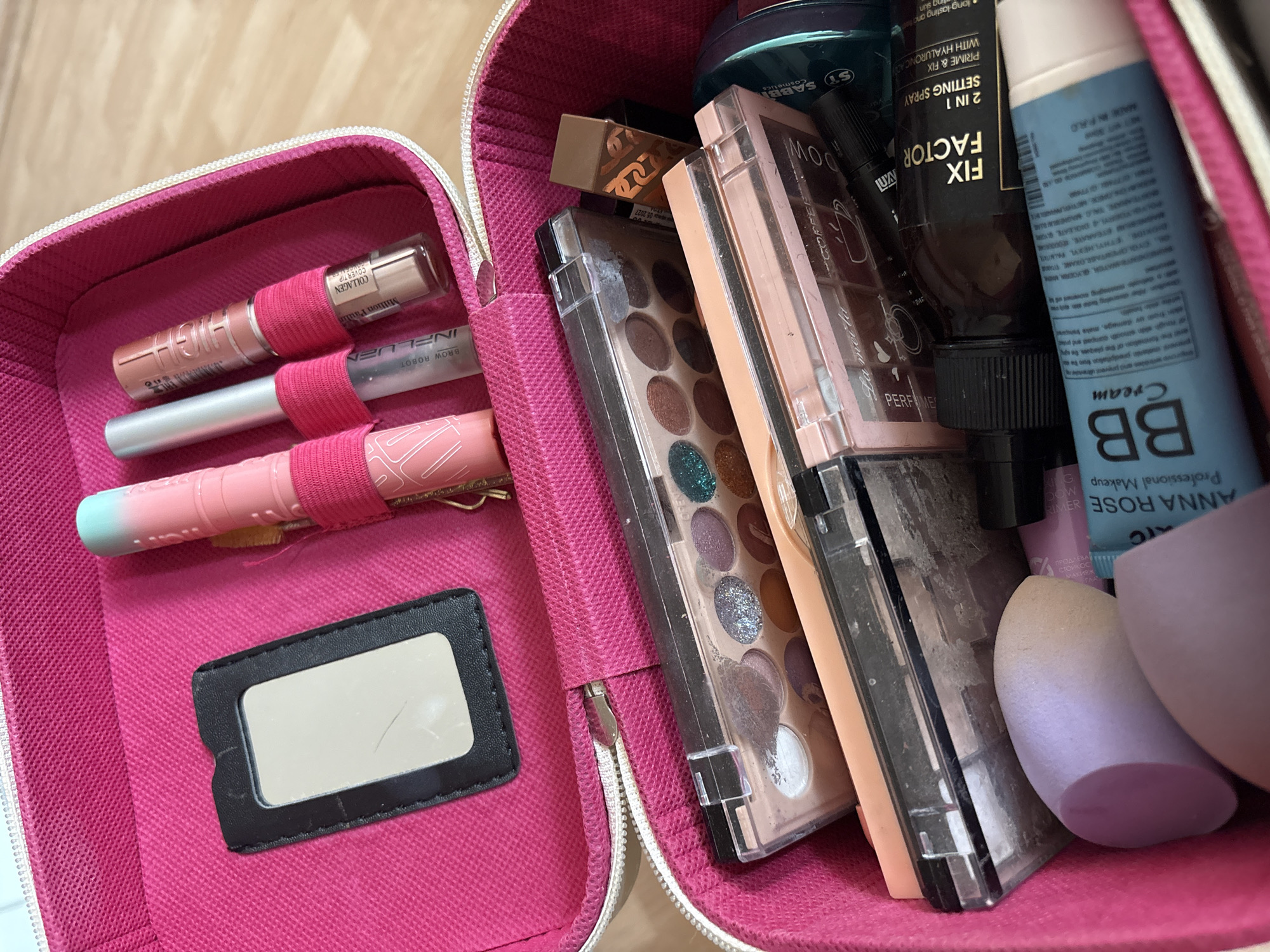 The image shows an open pink makeup bag. Inside, various makeup items are visible, including eyeshadow palettes, tubes of lip products, beauty sponges, and bottles of cosmetic products. Specifically, one product is labeled "ANNA ROSE Professional Makeup BB Cream". Another bottle indicates "FIX FACTOR 2 IN 1 PRIME & FIX SETTING SPRAY WITH HYALURONICAD". One of the items is also marked "BROW ROBOT INFLUZ". The makeup bag is filled to capacity. There is no location information provided in the image, nor is there any mention of people or actions taking place in Izhevsk, Russia.