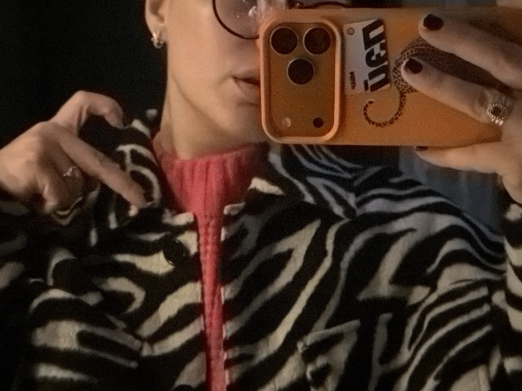 This is a close-up mirror selfie, likely taken indoors in Большая Шильна, Russia, featuring a partially visible person. The subject is wearing a vibrant pink turtleneck or sweater under a black and white zebra print jacket, which appears to have a plush or fuzzy texture.
The person's face is mostly obscured by an orange or peach-colored smartphone, which they hold with their right hand. The phone case clearly shows three circular camera lenses and two smaller circular details. On the back of the phone case, a sticker or design features the letters "USD" with "WRD" below it, along with a distinctive coiled snake or serpent pattern. The person's right hand has dark nail polish and wears a prominent, intricately designed ring on the ring finger.
With their left hand, the subject forms a heart shape with their fingers, visible against a dark background. A silver-toned ring is worn on the index finger of this hand. Part of the person's lower face, including their lips and chin, is visible, along with an ear adorned with a small silver hoop earring and the frame of a pair of glasses reflecting light. The background is dark and out of focus, suggesting an indoor setting with soft, slightly dim lighting. The overall image captures a casual and expressive moment.