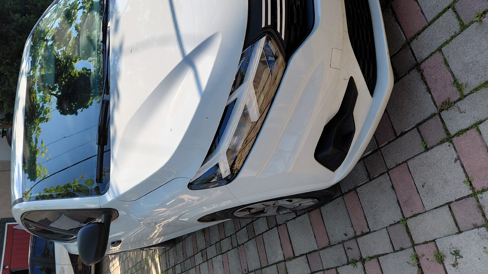 A white car is parked on a paver driveway outdoors. The car is viewed from the front quarter, with the driver's side front fender, headlight, and part of the hood visible. The car has a black side mirror and a black grille. The windshield reflects trees and the sky. The driveway is made of interlocking pavers in shades of gray and pink, with some weeds growing between them. The background shows a building with a red door and another vehicle. The scene is lit by natural sunlight, casting shadows on the car and driveway.