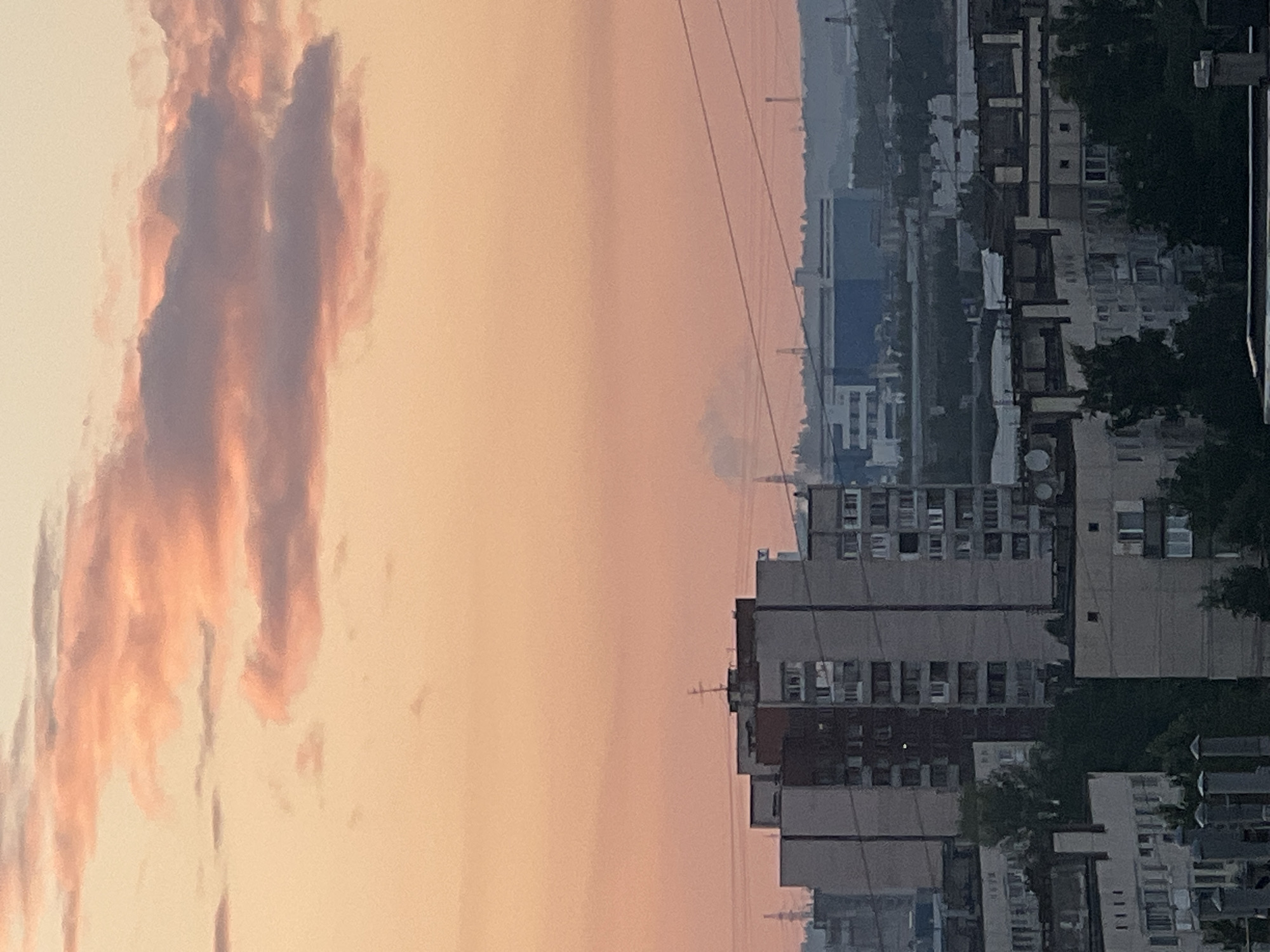 This media file captures a serene sunset over an urban landscape, likely Ladozhskoe Ozero, Russia. The sky is painted in hues of soft peach and pink, with wispy clouds that catch the fading light. These clouds, tinged with lavender and orange, dominate the left side of the frame, creating a dramatic contrast with the right side of the image.
The right side of the image presents a view of apartment buildings, their concrete facades catching the muted twilight. The buildings are densely packed, with visible windows and balconies suggesting a residential area. Trees, dark and silhouetted against the sky, are interspersed among the structures, adding a touch of nature to the urban scene. Power lines stretch across the frame, a common element in cityscapes.
The overall impression is one of tranquility and the quiet transition from day to night. The time of day is clearly sunset, with the warm, diffused light characteristic of this time. The weather appears clear, allowing for the unobstructed view of the sunset and the cityscape. There is no visible text in the image. The scene evokes a sense of calm and the everyday beauty of a city at dusk.