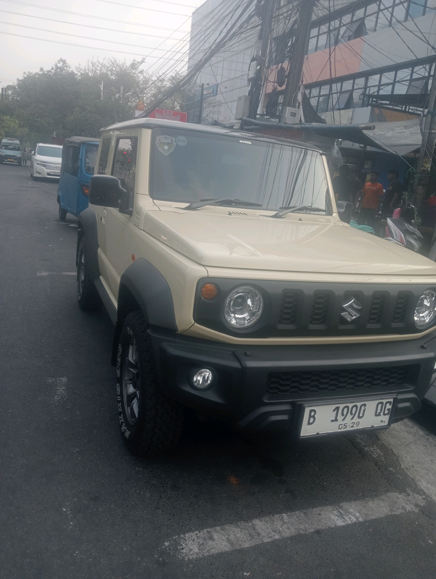 An outdoor, slightly hazy daytime, street-level medium shot captures a cream-colored Suzuki Jimny parked on an asphalt road. The vehicle is the primary subject, positioned at a slight angle, with its front end facing right. Its headlights, grille, and front bumper are visible. A license plate reading "B 1990 QG 05.29" is affixed to the front bumper. In the background, other vehicles, including a blue three-wheeled vehicle and a white car, are also present. A building with a glass facade and a complex network of overhead electrical wires dominates the upper right portion of the frame. The overall environment suggests a commercial or urban setting within East Jakarta, Indonesia.