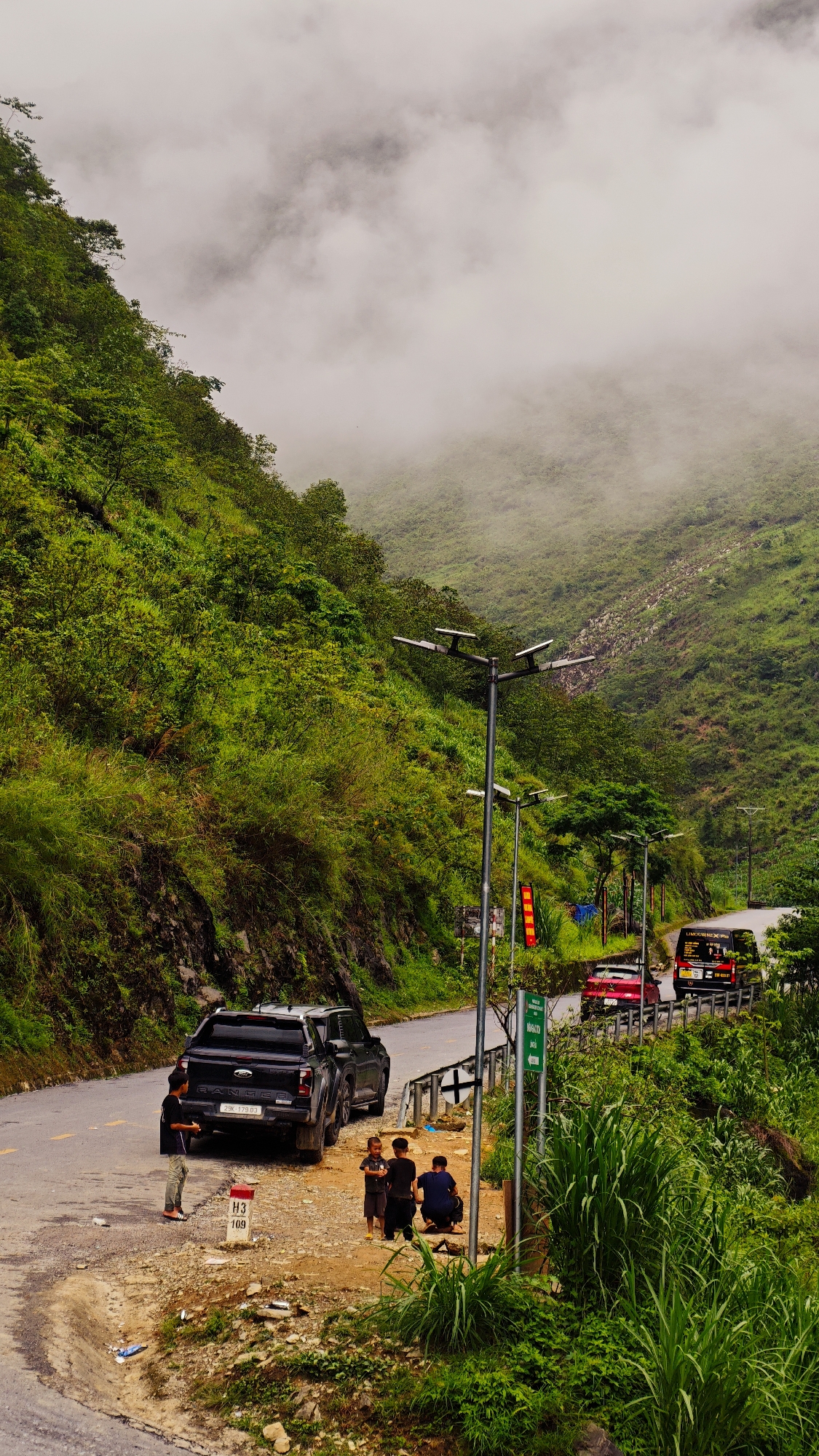 Captured in Yên Minh Commune, Vietnam, a narrow road winds through a lush, mountainous landscape shrouded in mist. The scene reveals a black Ford Ranger pickup truck parked on the roadside, its license plate reading "29K 179.03." Nearby, four children are gathered by a signpost marked "H3 109." Further down the road, a red car and a black van are visible. The roadside features green vegetation and utility poles. The backdrop consists of verdant hills and fog.