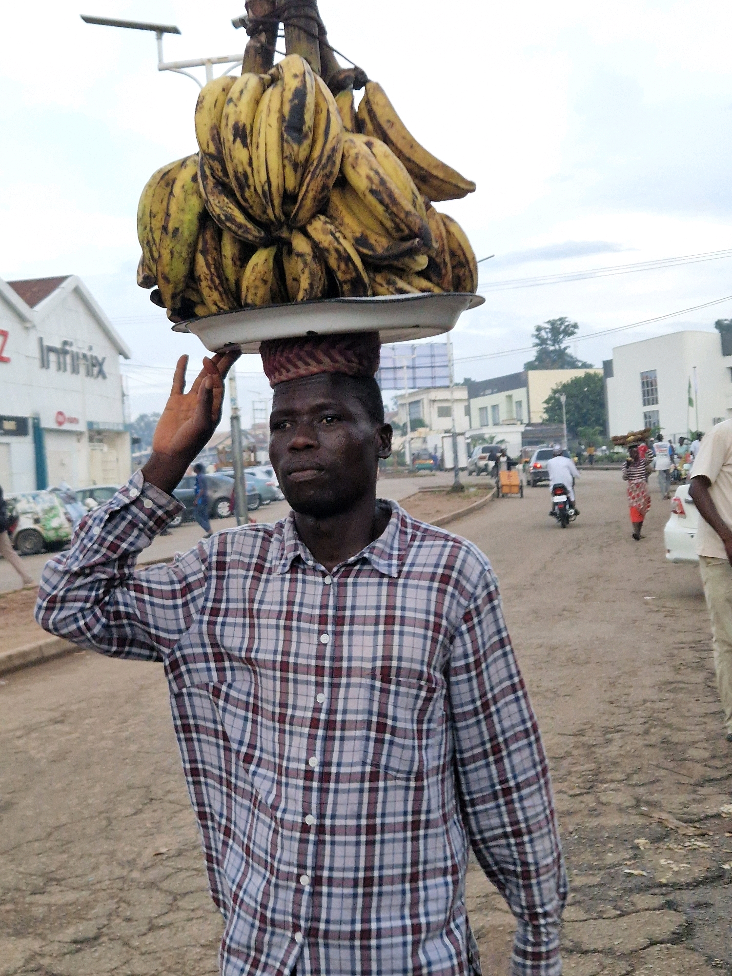 A man with dark skin, wearing a red, white, and blue plaid shirt, balances a large bunch of yellow bananas on his head. He is walking on a paved road in a city. The road is cracked and uneven, with debris scattered around. In the background, there are buildings, vehicles, and other people. To the left, there is a white building with a blue awning and the word "Infinix" written on it. To the right, there are more buildings and trees. The sky is overcast. The scene appears to be a street market or a busy urban area.