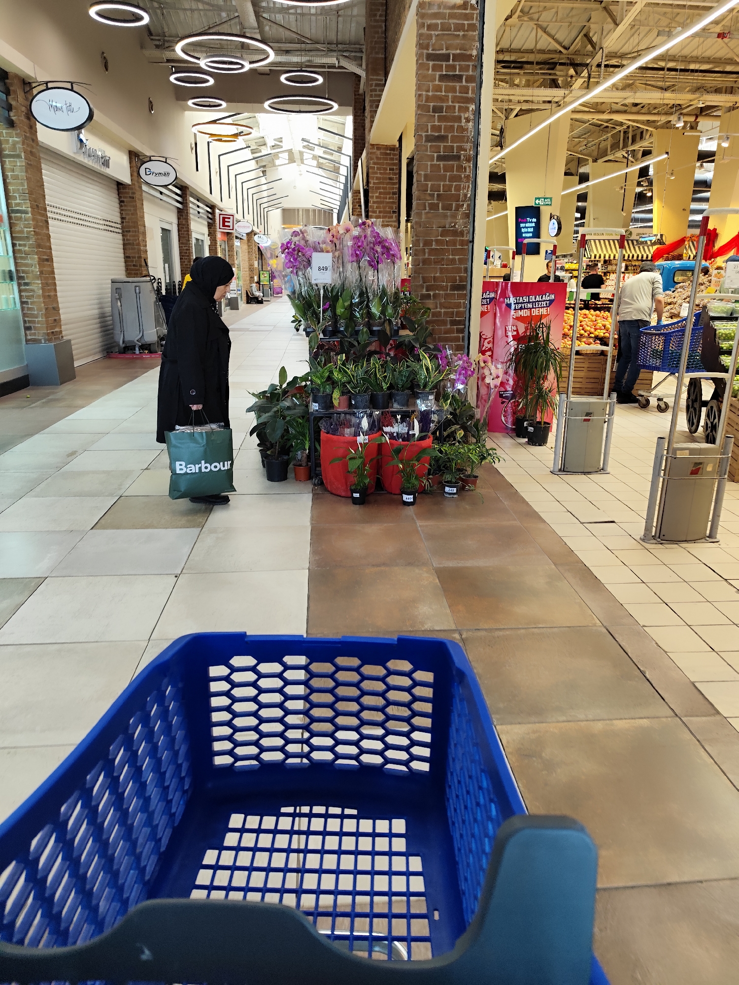 The image shows the inside of a shopping mall in Sarıyer, Turkey. A woman wearing a black abaya and hijab is carrying a green "Barbour" shopping bag. She is standing near a display of potted plants with pink orchids. A blue plastic shopping cart is in the foreground. In the background, people are shopping in a brightly lit supermarket. Signs are visible in Turkish.