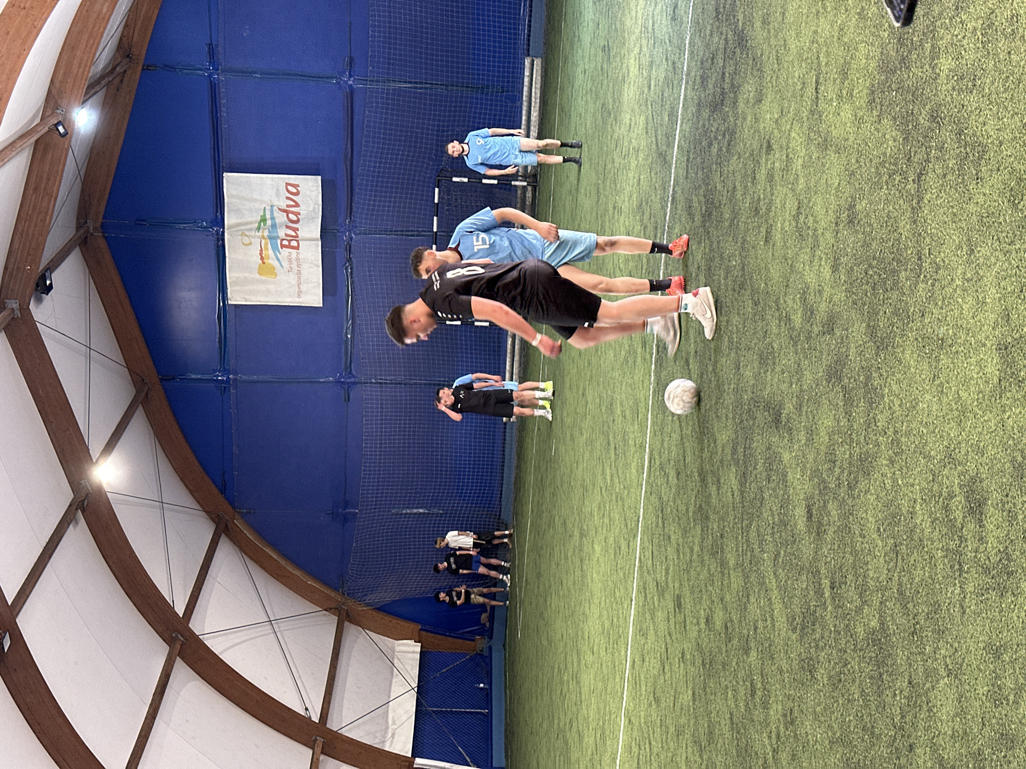 The image depicts an indoor soccer game or practice session in progress. The scene is set on an artificial turf field within a domed structure with exposed wooden support beams. A banner with "Budva" displayed on it hangs on the netting behind the goal, suggesting the location is in Budva, Montenegro. Several players, wearing light blue and black uniforms, are standing on the field. One player in black appears to be in motion, possibly kicking a soccer ball positioned near him. A group of people are observing from the sidelines. The overall impression is of an active indoor sports facility.