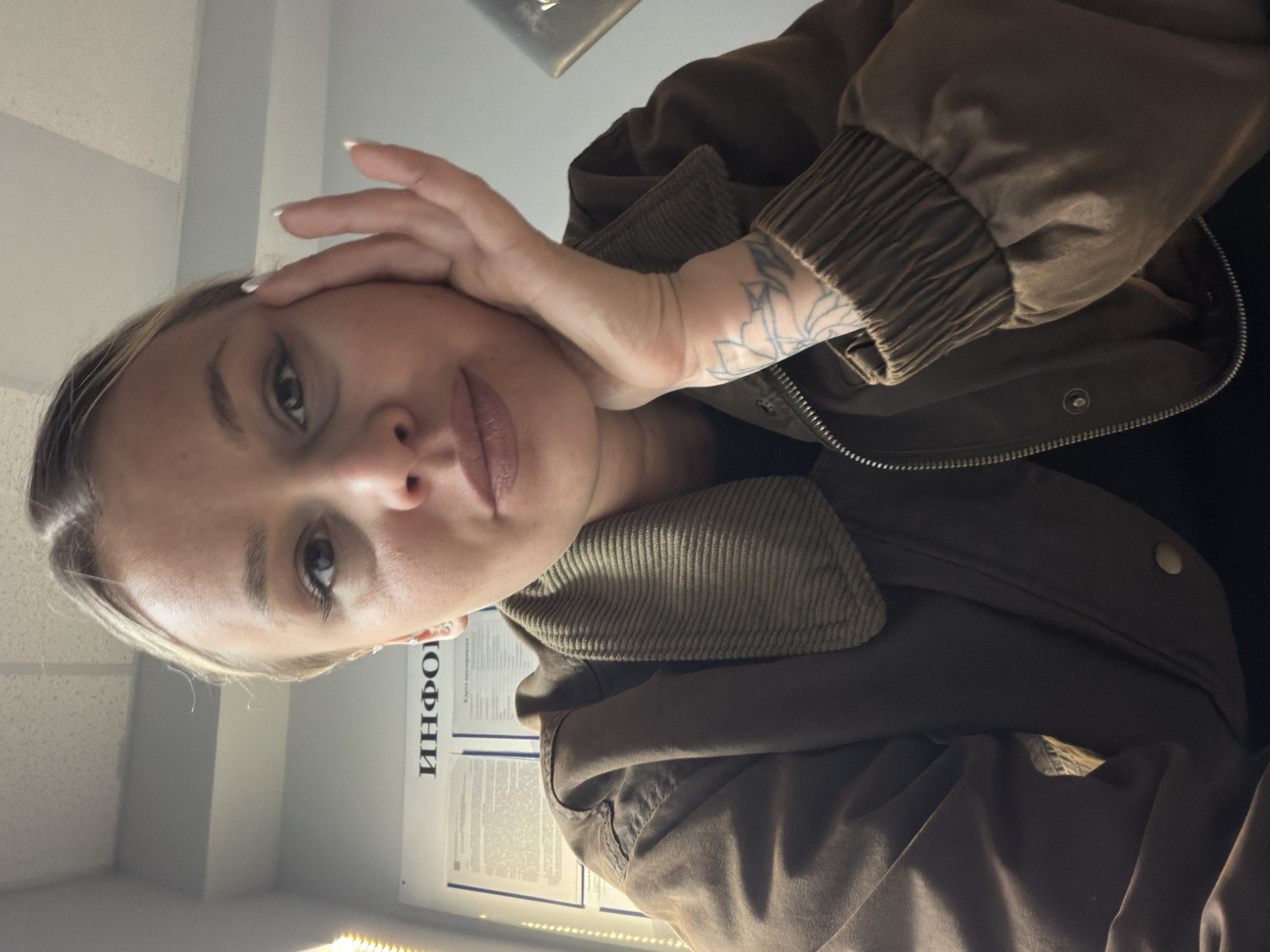 A woman with fair skin and blonde hair, wearing a brown jacket, is posing for a close-up selfie. She is looking at the camera with a neutral expression. Her right hand is raised to her forehead. A visible tattoo is on her left wrist. Behind her is a poster with Russian text that includes the word "ИНФОРМАЦИЯ" (Information) and a bright light. The location appears to be an indoor space, possibly in Sarapul, Russia, based on the Cyrillic text.