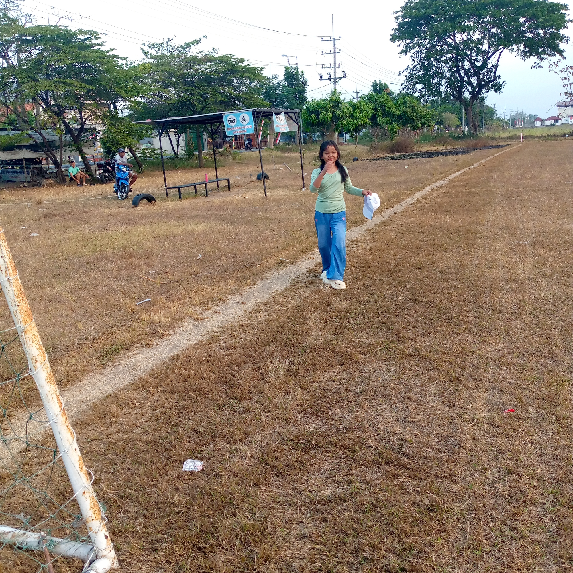 The image depicts an outdoor scene in the city of Pacing, Indonesia. A young girl with dark hair, wearing a light green long-sleeved shirt and blue jeans, is walking on a dry, grassy field. She is holding a white cap in her right hand and appears to be looking towards the camera, with her mouth slightly open. The field has a white line marking a path, and beyond it, a covered shelter with benches and a motorcycle is visible. In the background, trees and power lines can be seen, along with some distant buildings. The overall environment appears somewhat neglected, with dry vegetation and scattered debris. To the left, a rusty white goalpost with netting suggests the field might be a sports ground.