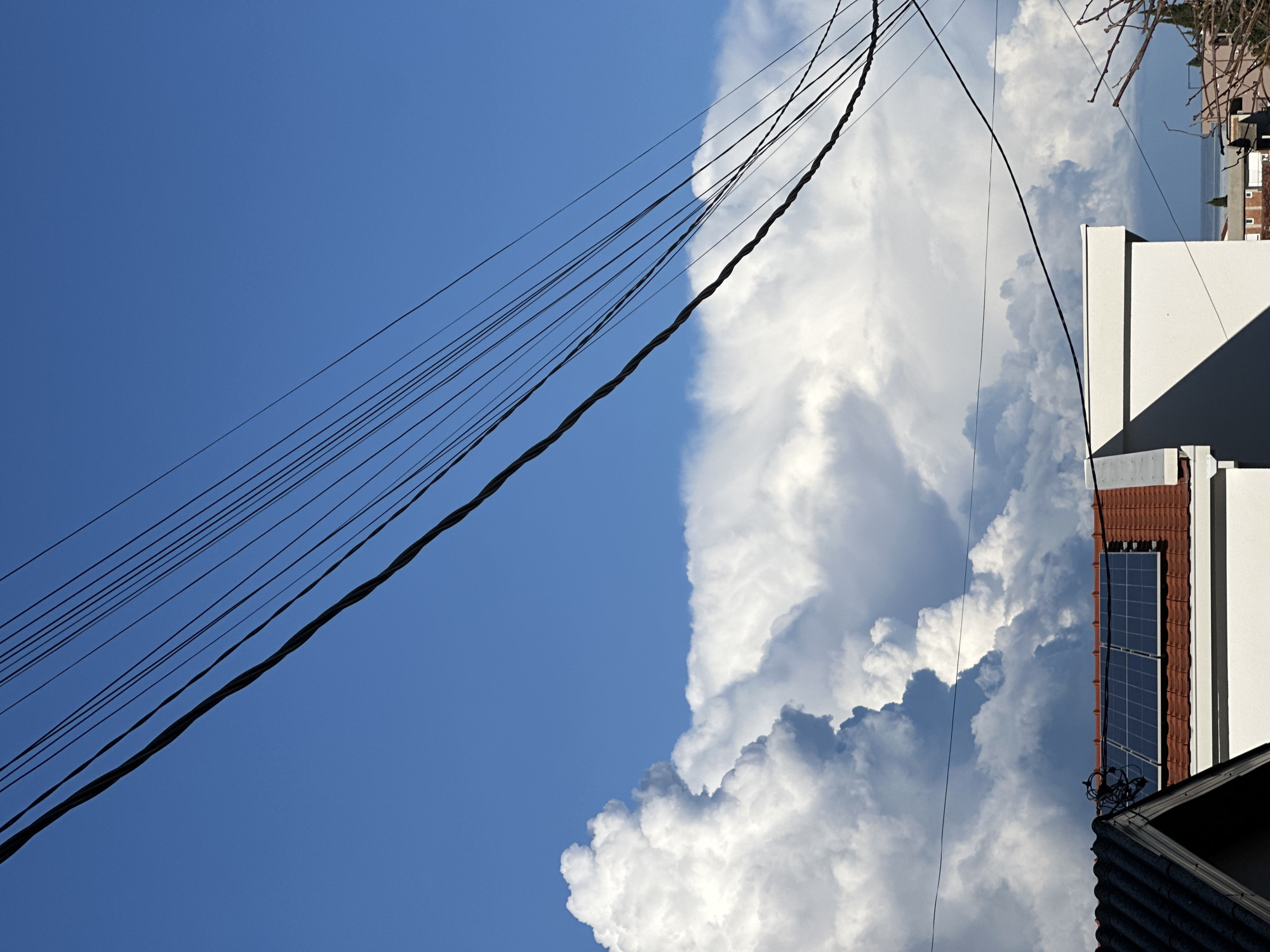 The image presents an outdoor scene captured in Šušanj, Montenegro, dominated by sky and urban elements. The upper-left quadrant of the frame features a clear, deep blue sky. Below and to the right, a large formation of dense, white cumulus clouds with some grey undersides extends across the mid-ground. Multiple black utility lines, comprising two thicker braided cables and several thinner parallel wires, traverse the upper half of the image diagonally from the upper left to the lower right, crossing both clear sky and clouds.
In the lower left, a portion of a building with a dark grey tiled gable roof is visible. Adjacent to it, towards the center-right, stands a light-colored building with a red tiled roof. Mounted on this red roof is an array of three dark-blue rectangular solar panels. A distinct shadow is cast on the side of the light-colored building, indicating direct sunlight from the upper left. In the distant background on the right side, a segment of a calm blue sea is visible above muted structures and partially obscured by bare tree branches. No individuals or specific actions are discernible.
