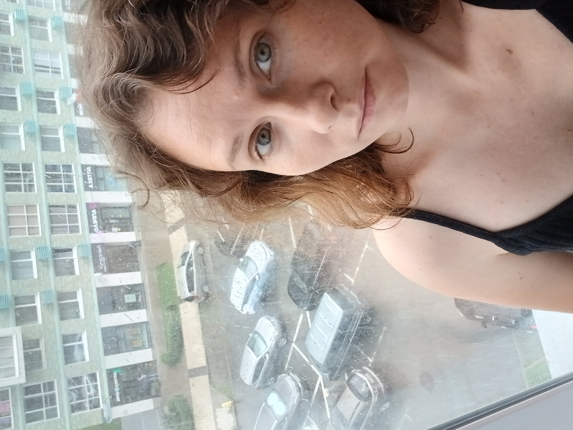 A selfie of a young woman with curly, light brown hair and light blue eyes, taken indoors looking out a window on a rainy day. The woman is positioned in the lower right quadrant of the frame, looking directly at the camera. Her expression is neutral. She is wearing a black camisole.
Through the window, a building with multiple floors and windows is visible. Below the building, several cars are parked in a lot. The rain on the window creates streaks and blurs, affecting the clarity of the outdoor scene. The architecture of the building and the presence of a Russian flag sticker on one of the shops visible through the window suggest the location is Yekaterinburg, Russia. Some of the shops visible through the window have Cyrillic signage, further indicating a Russian location.