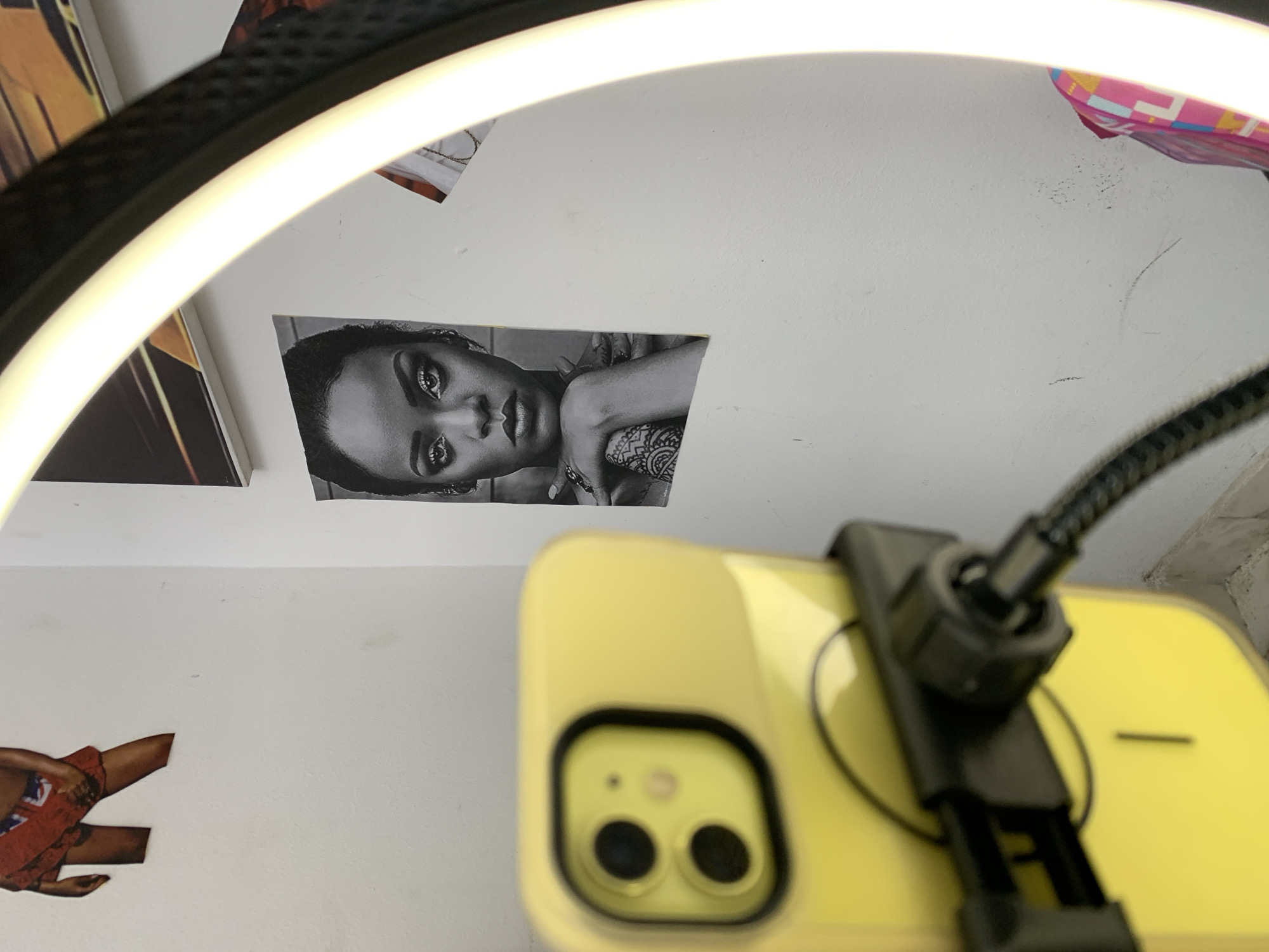 A close-up, low-angle shot captures a yellow smartphone case held by a black mount, with a black cable connected to it, situated in what appears to be a room in Lagos, Nigeria. The background features a white wall adorned with several posters. One poster, a black and white image, depicts the face of a woman with dramatic eye makeup and intricate tattoos on her arm. Another poster shows a fragment of a person's lower body, wearing a red skirt with a white and blue pattern. A brightly lit ring light is partially visible in the foreground on the right side of the frame, casting a strong glow. The overall impression is that of a setup for recording content, possibly a vlogging or social media setup, in a personal space.