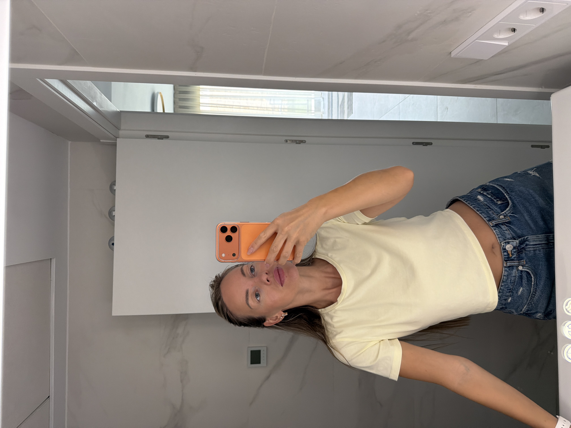 A young woman with long brown hair is taking a mirror selfie in what appears to be a bathroom, likely located in Finestrat, Spain. She is wearing a light yellow crop top and distressed denim shorts. She is holding an orange smartphone in her right hand, angled to capture her reflection. The background shows a modern bathroom with marble-patterned walls and a white door. In the reflection of the mirror, a doorway leads to another room with sheer curtains and natural light. Electrical outlets are visible on the right-hand wall.