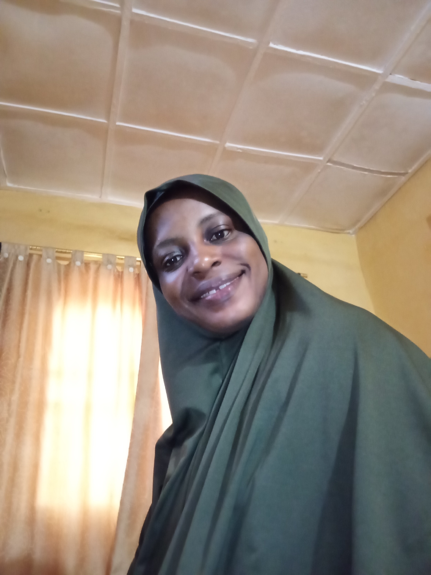 A woman in a green hijab smiles at the camera. She is indoors, with a beige ceiling and yellow walls visible. Behind her, on the left, are beige curtains that are illuminated from behind by a bright light. The image appears to be a selfie. The location is identified as Narayi, Nigeria.