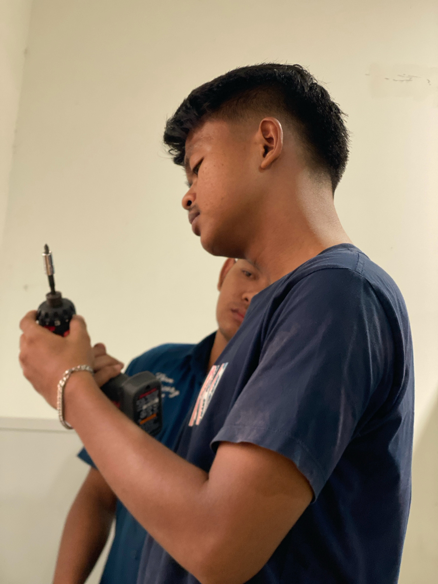In Batam, Indonesia, a young man with a dark buzz cut and a short fade haircut is shown in profile, holding a black and red power drill with a silver bit. He wears a navy blue t-shirt and a silver bracelet on his left wrist. Another young man, also wearing a navy blue shirt, is partially visible in the background, looking forward. The background is a plain, light-colored wall, suggesting an indoor setting. The lighting appears to be artificial and evenly distributed. The image captures a moment of focused activity, likely involving some form of construction or repair work.