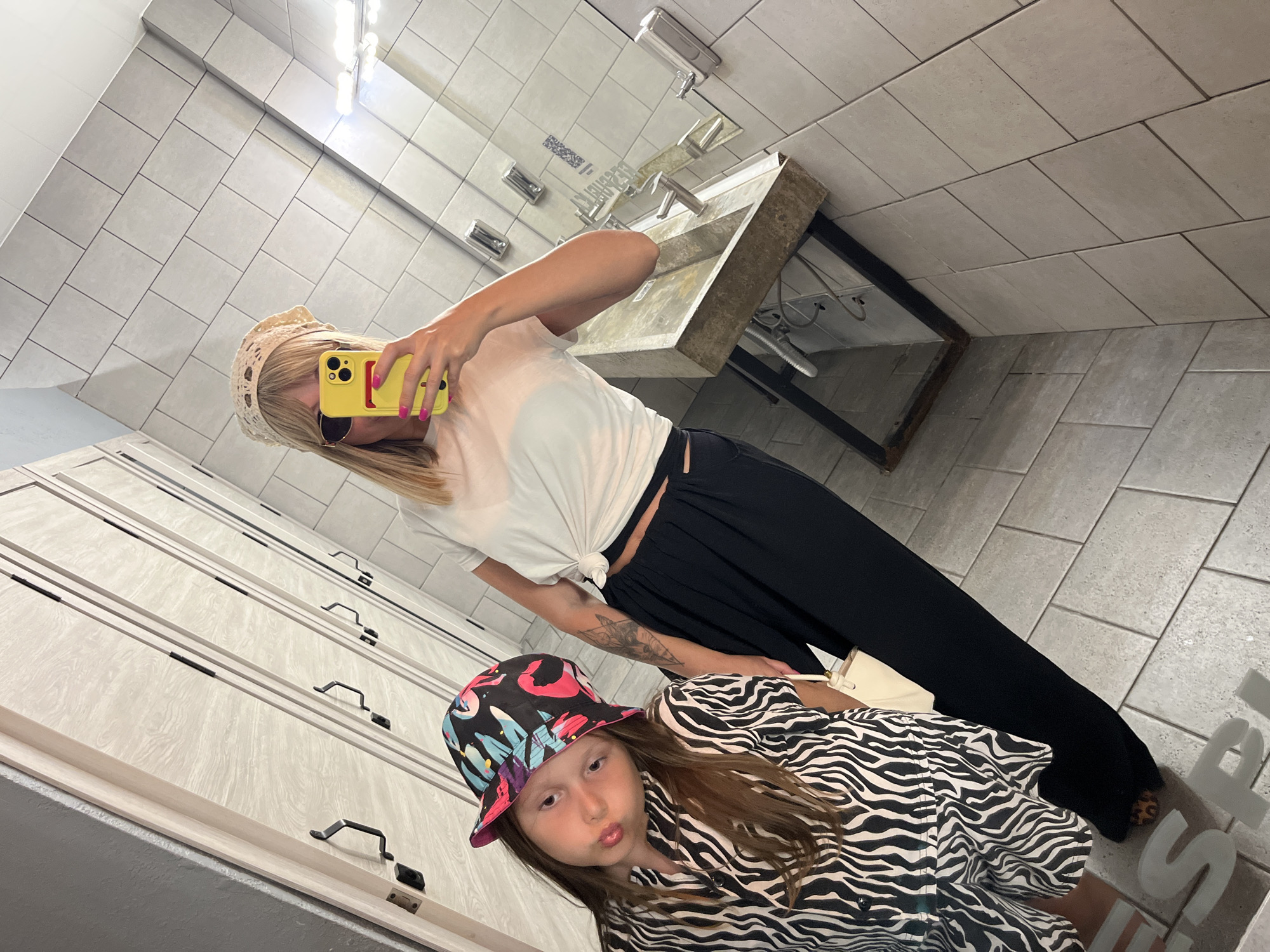 A woman and a young girl pose for a selfie in a brightly lit, tiled bathroom, likely in a public restroom. The woman, wearing a white t-shirt knotted at the waist, dark pants, sunglasses, and a cream-colored headscarf, holds up a yellow smartphone to capture the reflection. The girl, to her left, wears a black and white zebra-print shirt and a colorful bucket hat. She puckers her lips at the camera. Behind them, a concrete sink with chrome faucets is visible, along with mirrored walls and cabinetry. The floor is tiled in a light gray, and large white lettering is visible on the floor in the foreground.