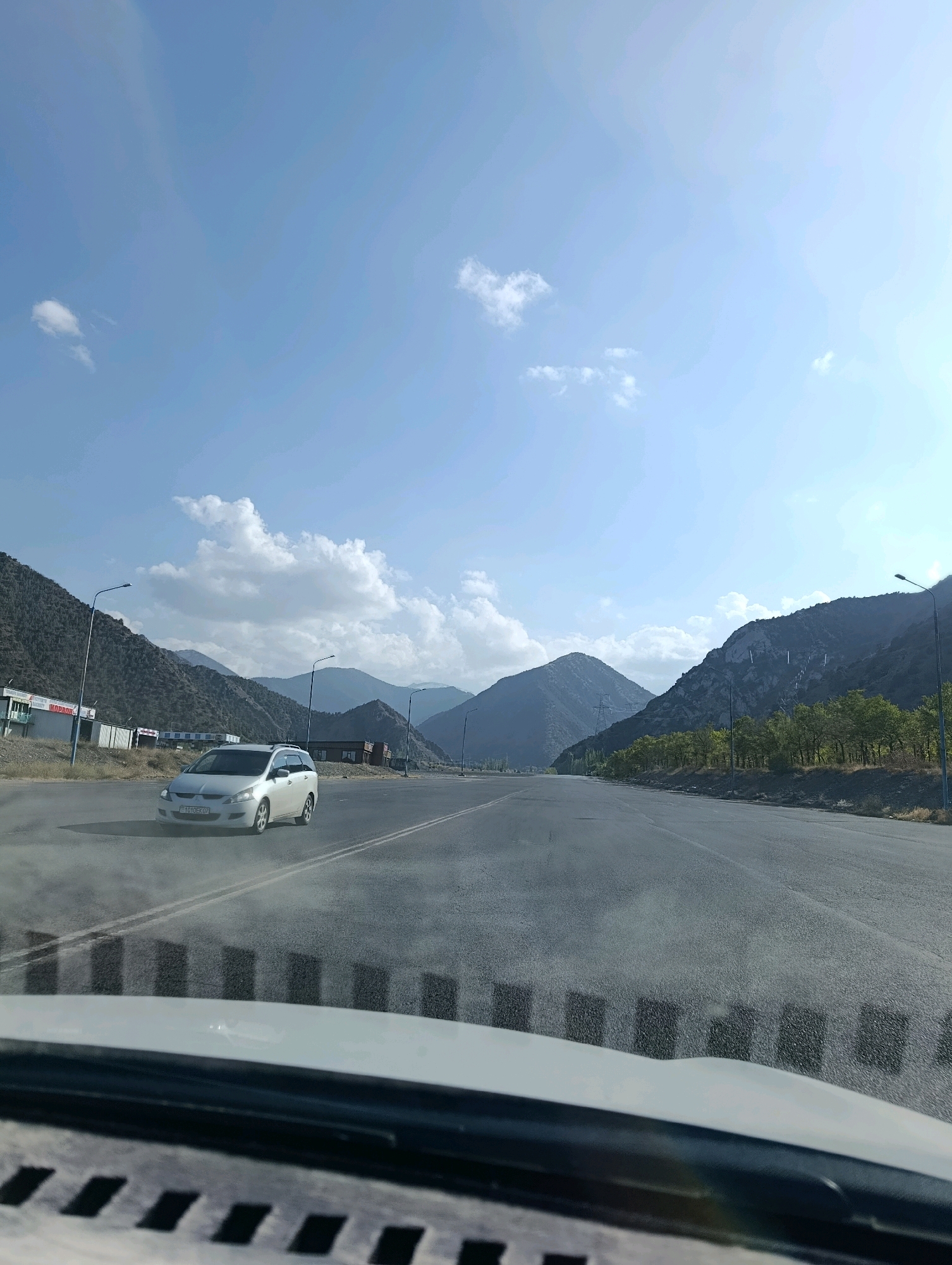 An eye-level shot captures a road winding through a mountainous landscape under a clear blue sky with scattered white clouds. A white minivan is driving on the right lane of the paved road, its headlights on. The road is flanked by steep, rocky mountains on both sides, sparsely covered with trees and vegetation. On the left side of the road, there are some low-rise buildings and streetlights. The foreground shows a portion of a car's interior dashboard, suggesting the photo was taken from inside a vehicle.