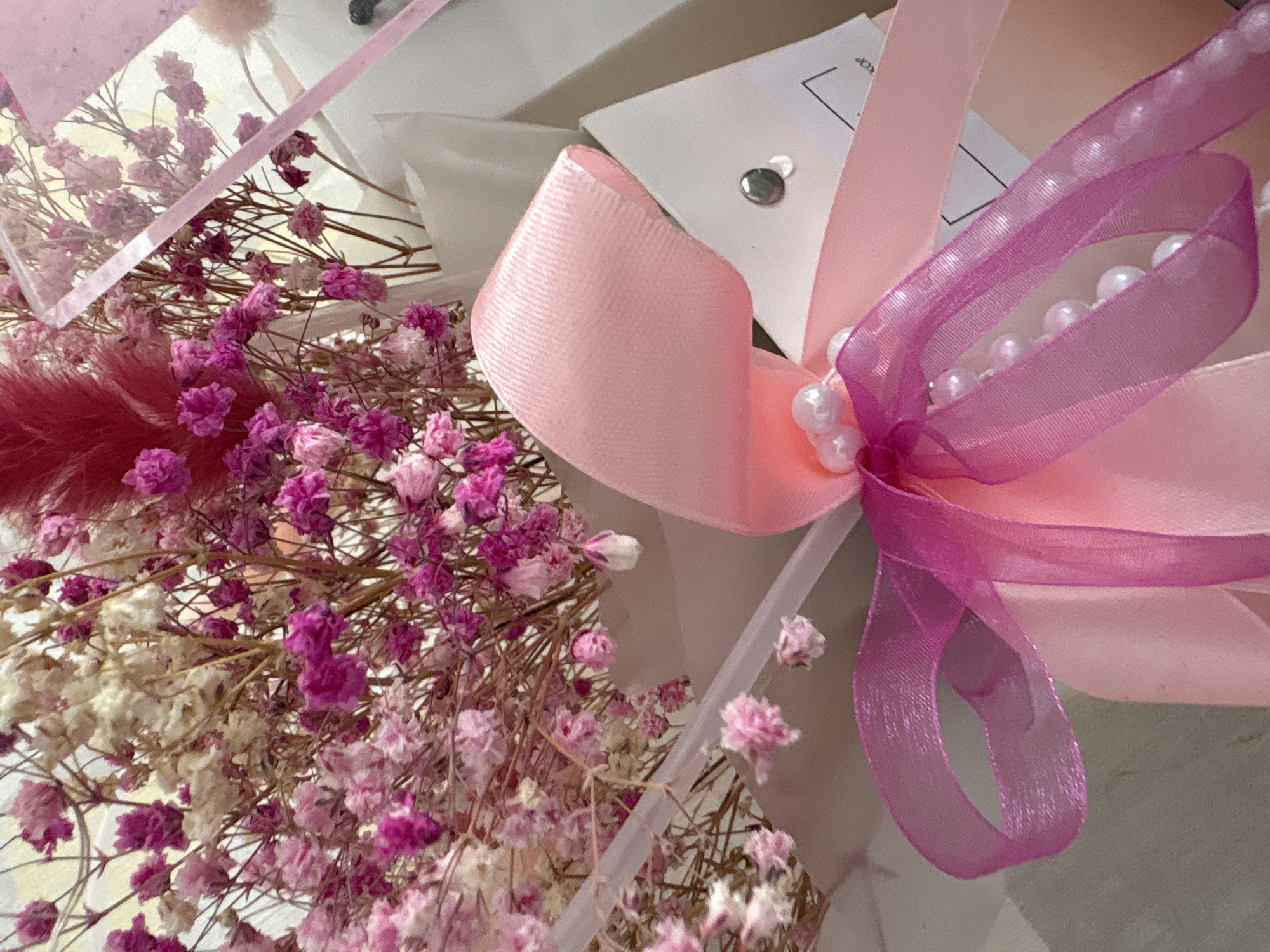 The image shows a close-up of a floral arrangement. Predominantly, there are various shades of pink and white dried flowers, including what appears to be baby's breath. The flowers are bundled together, and a pink ribbon with a lighter pink satin bow and a purple sheer ribbon with pearl beads are tied around them. A white card with an attachment is partially visible. The overall setting appears to be indoors, with soft lighting. The location is not evident.